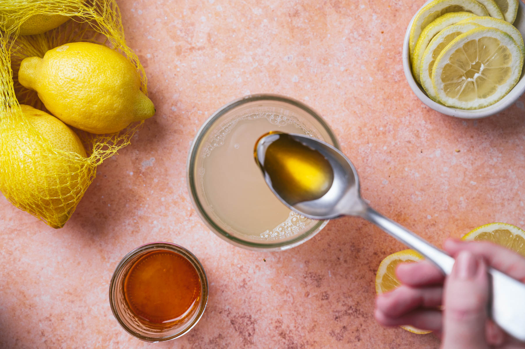 A spoonful of honey is being added to a glass of lemon water. Lemons in a net bag, lemon slices, and a jar of honey are placed on an orange countertop.