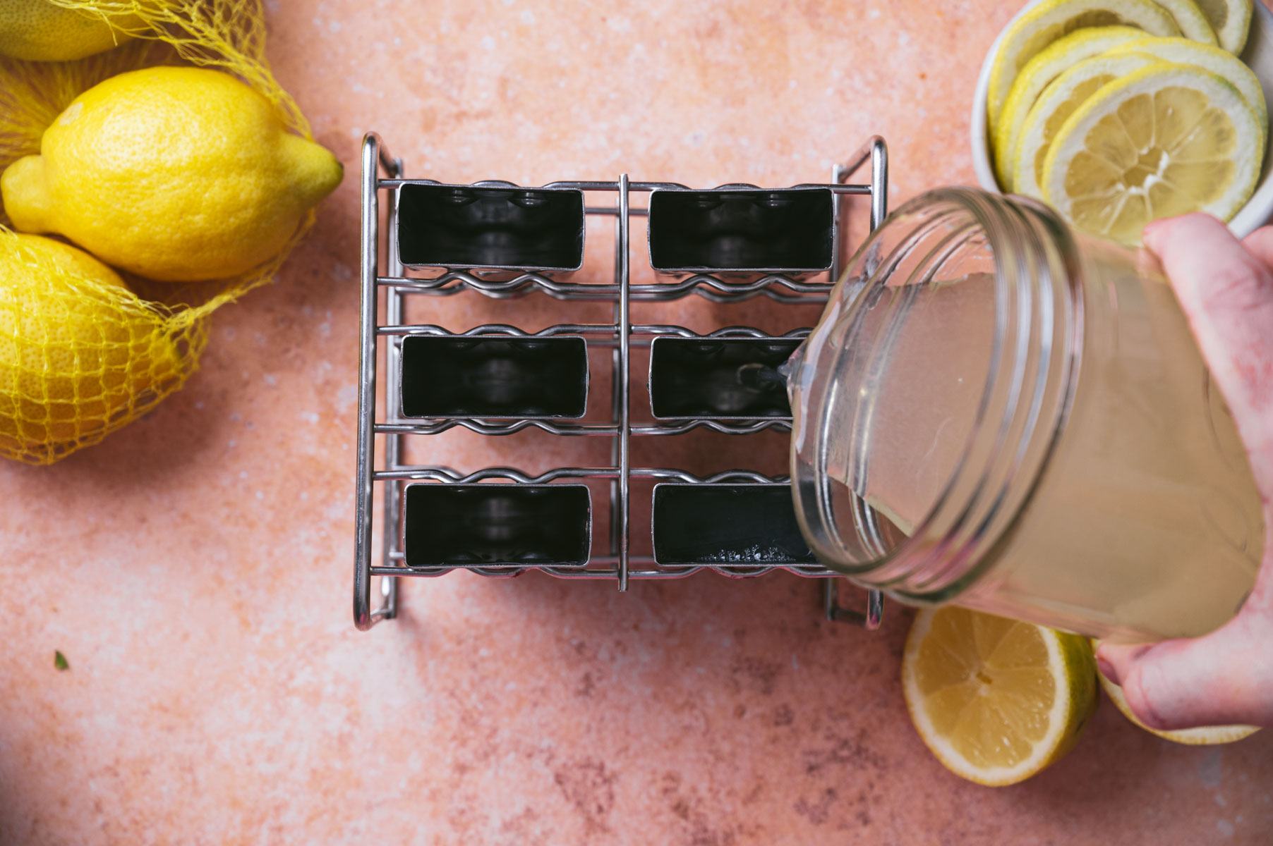 A person pours lemonade from a jar into six small glass slots in a metal rack on a countertop with lemons.
