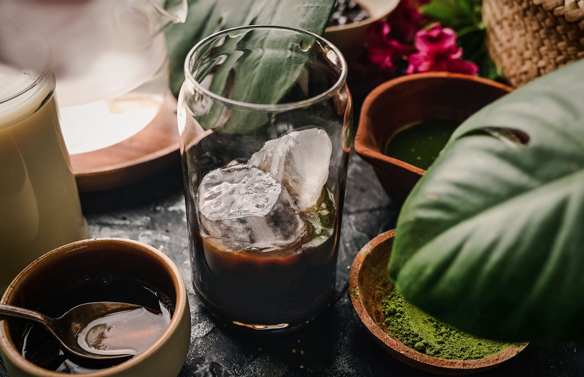 A glass of iced coffee is surrounded by bowls containing ingredients like matcha powder, a spoon in a dark liquid, and greenery, set on a dark surface.