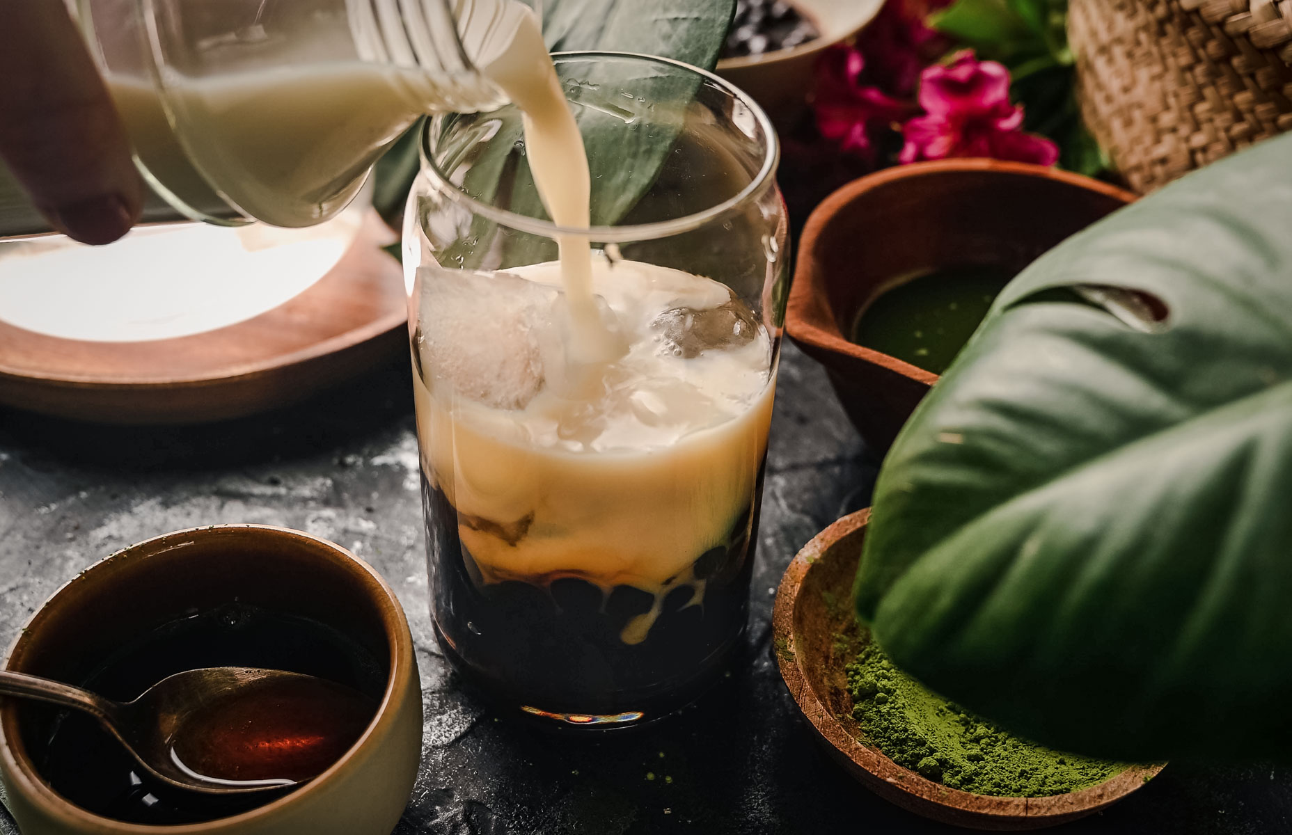 A close-up of a glass being filled with milk tea over ice. The glass contains dark tapioca pearls. Surrounding the glass are bowls of brown liquid, green powder, and leaves.