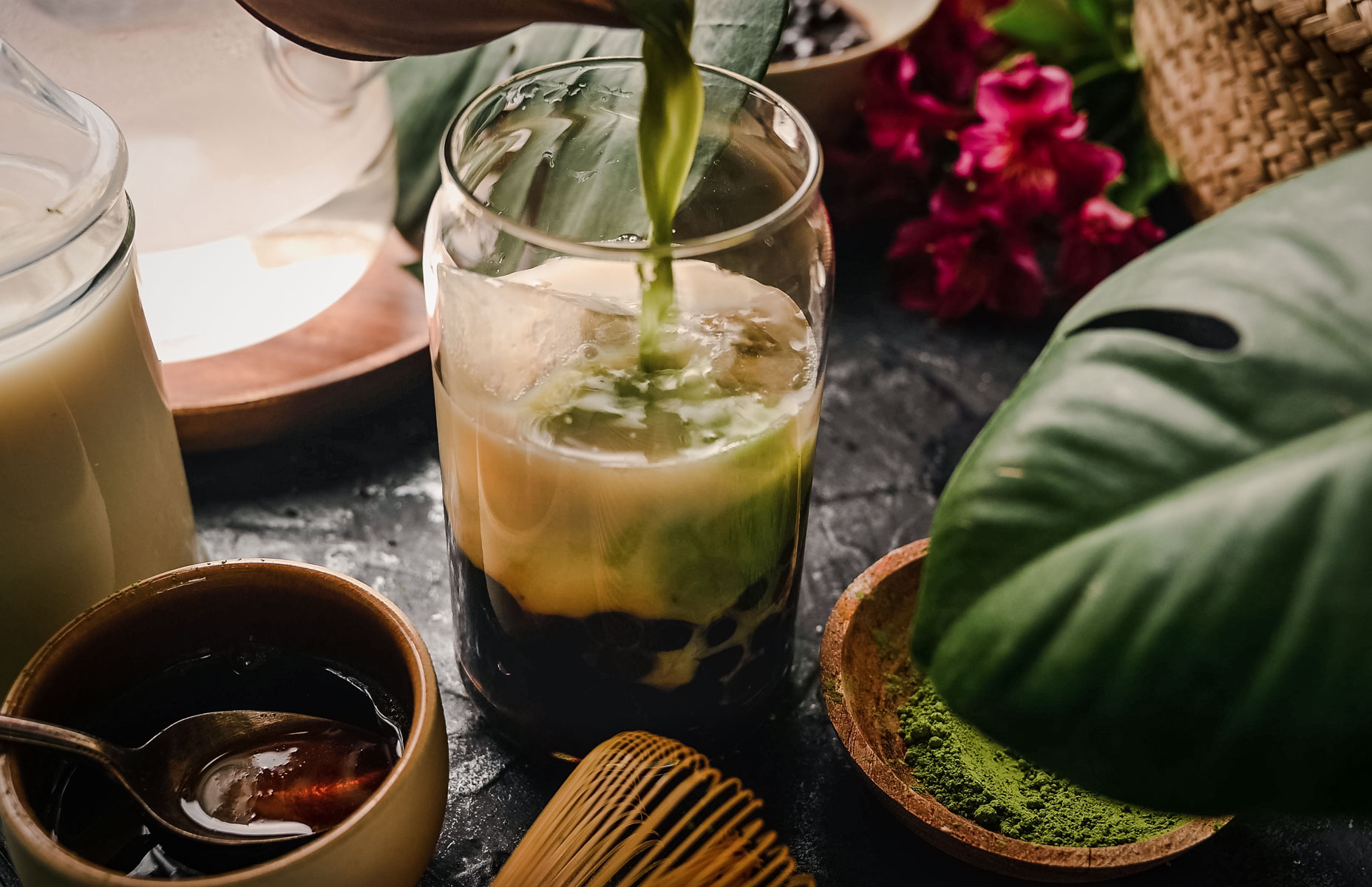 A glass being filled with green matcha tea over milk and tapioca pearls, surrounded by various ingredients and green leaves.