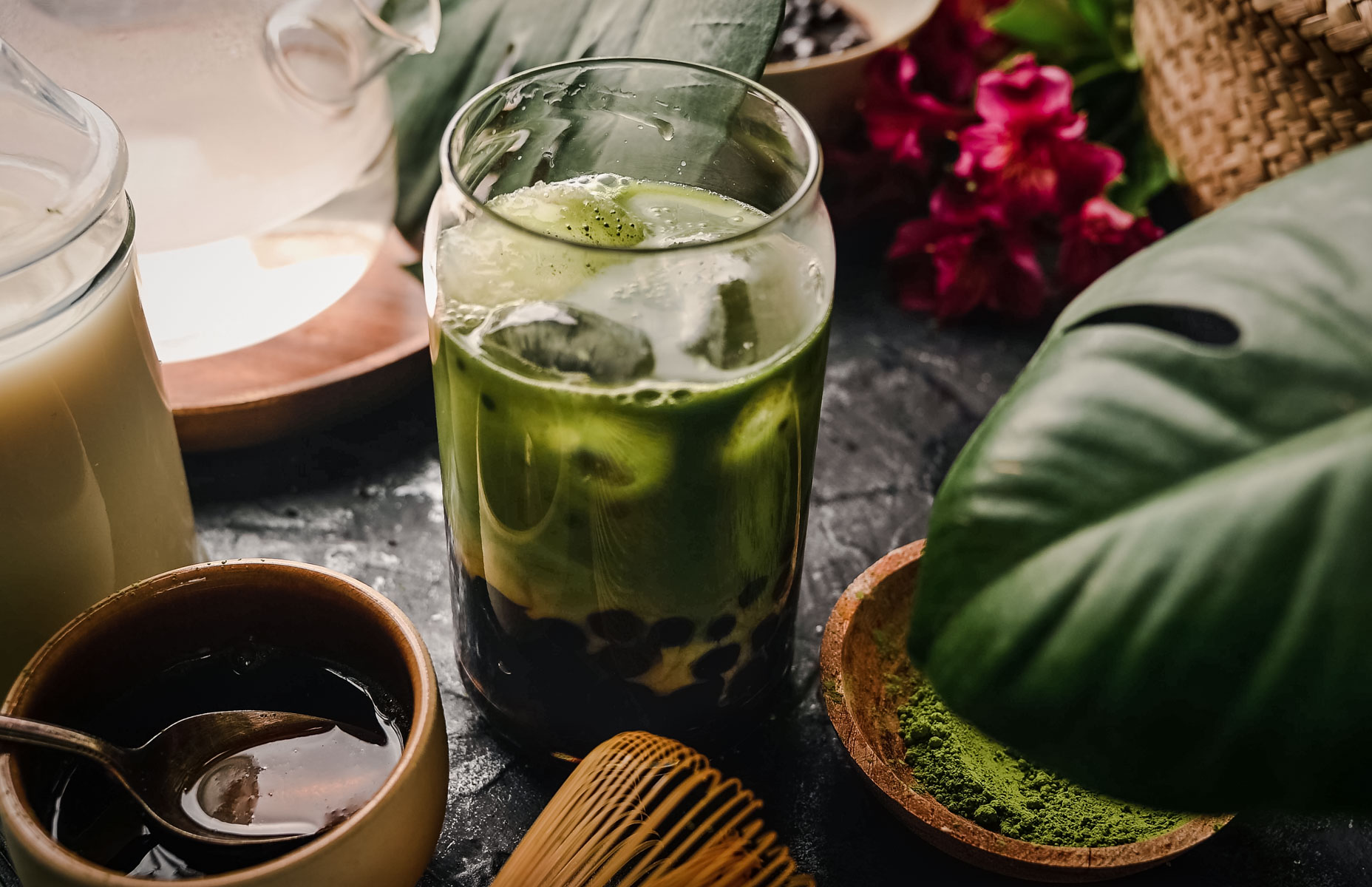 A glass of iced matcha green tea with tapioca pearls sits on a table surrounded by matcha powder, a bamboo whisk, and leaves.