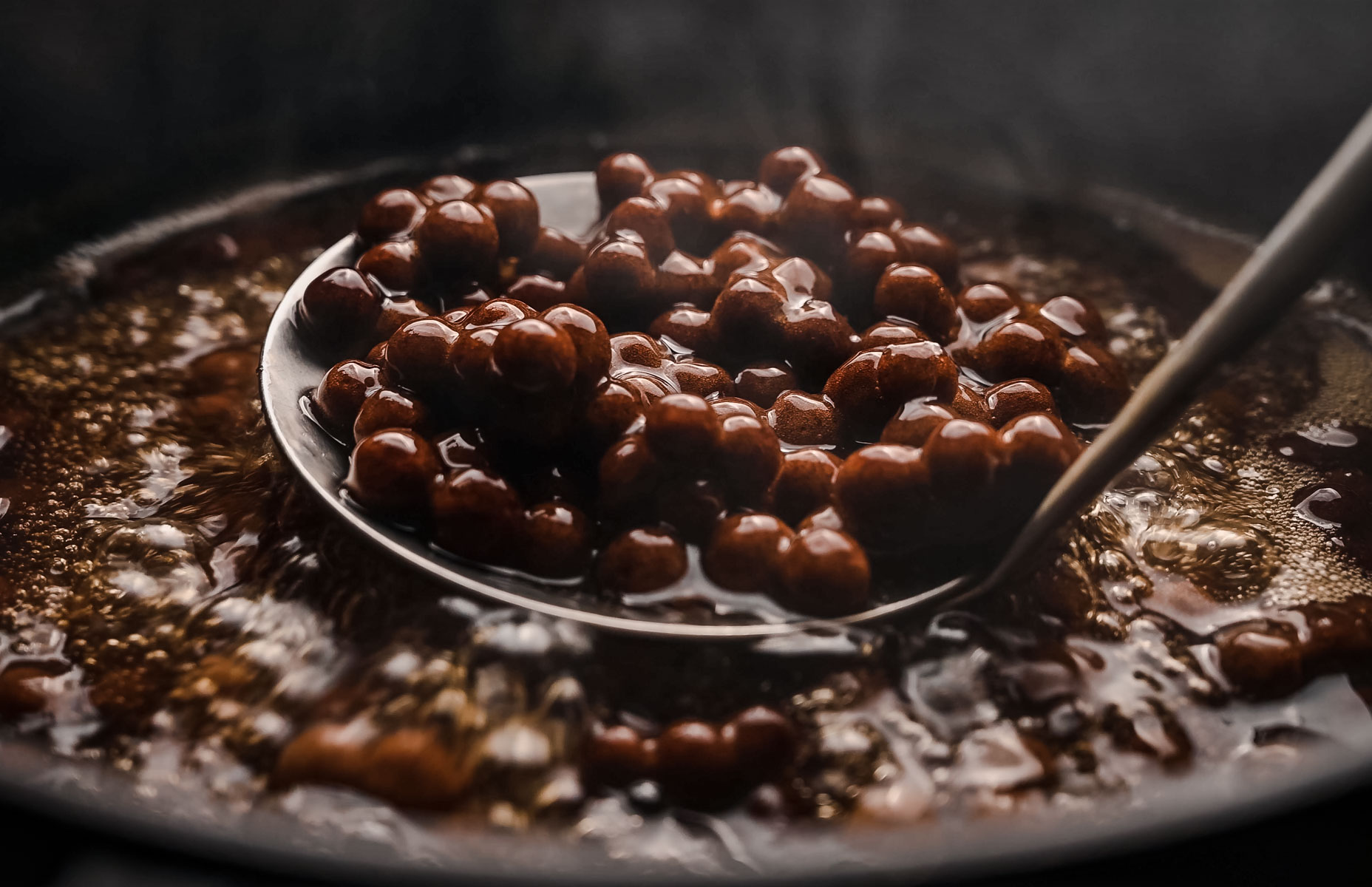 A ladle scooping brown tapioca pearls from a dark boiling liquid.
