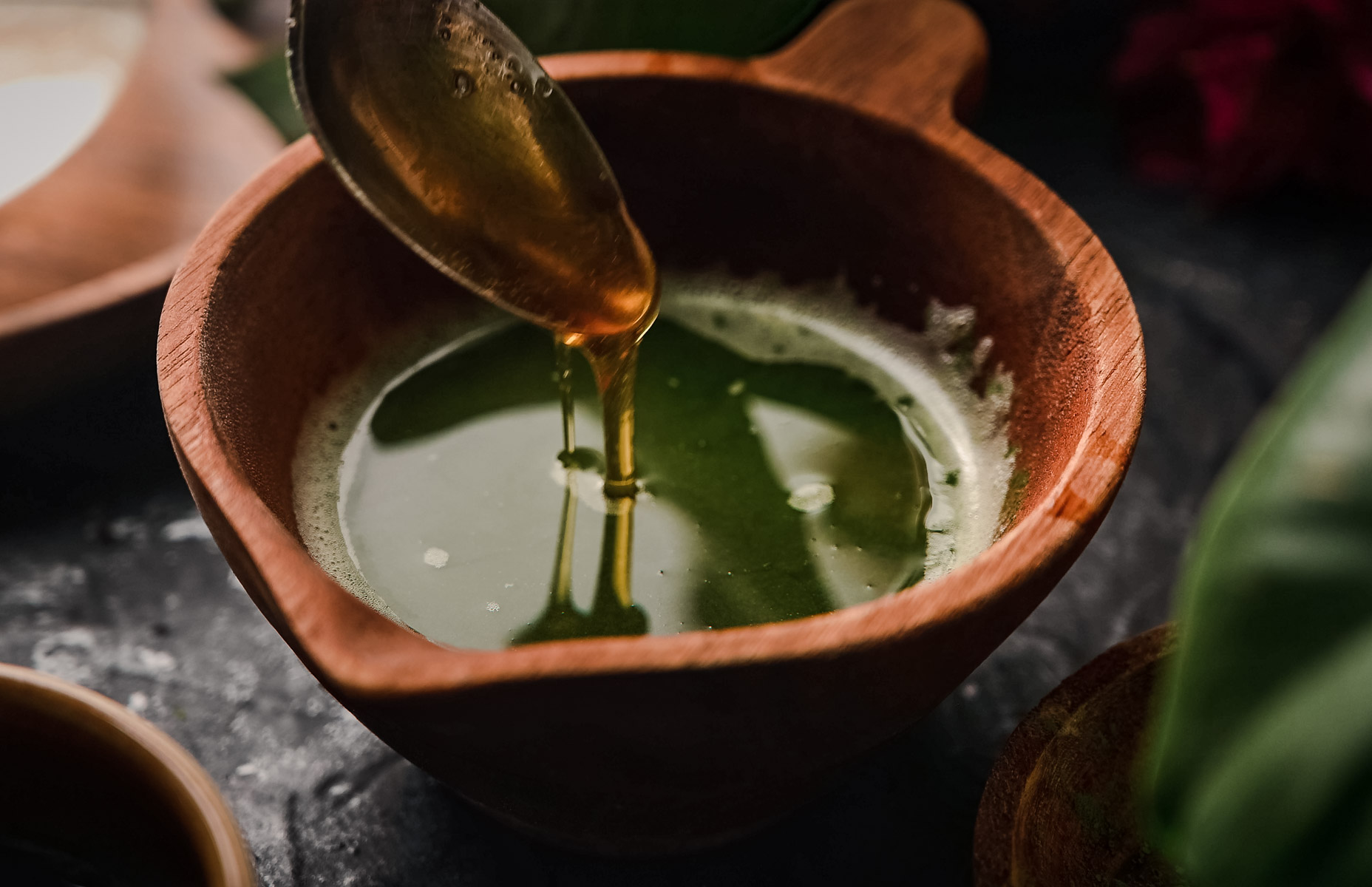 A spoon drizzles maple syrup into a wooden bowl containing green liquid.