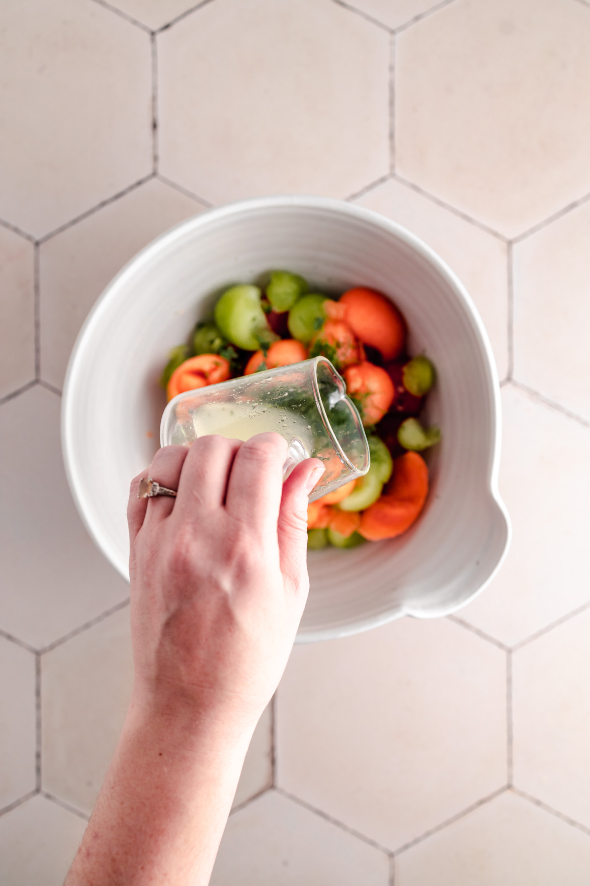 A hand pours liquid from a small glass into a large white bowl containing vegetables.
