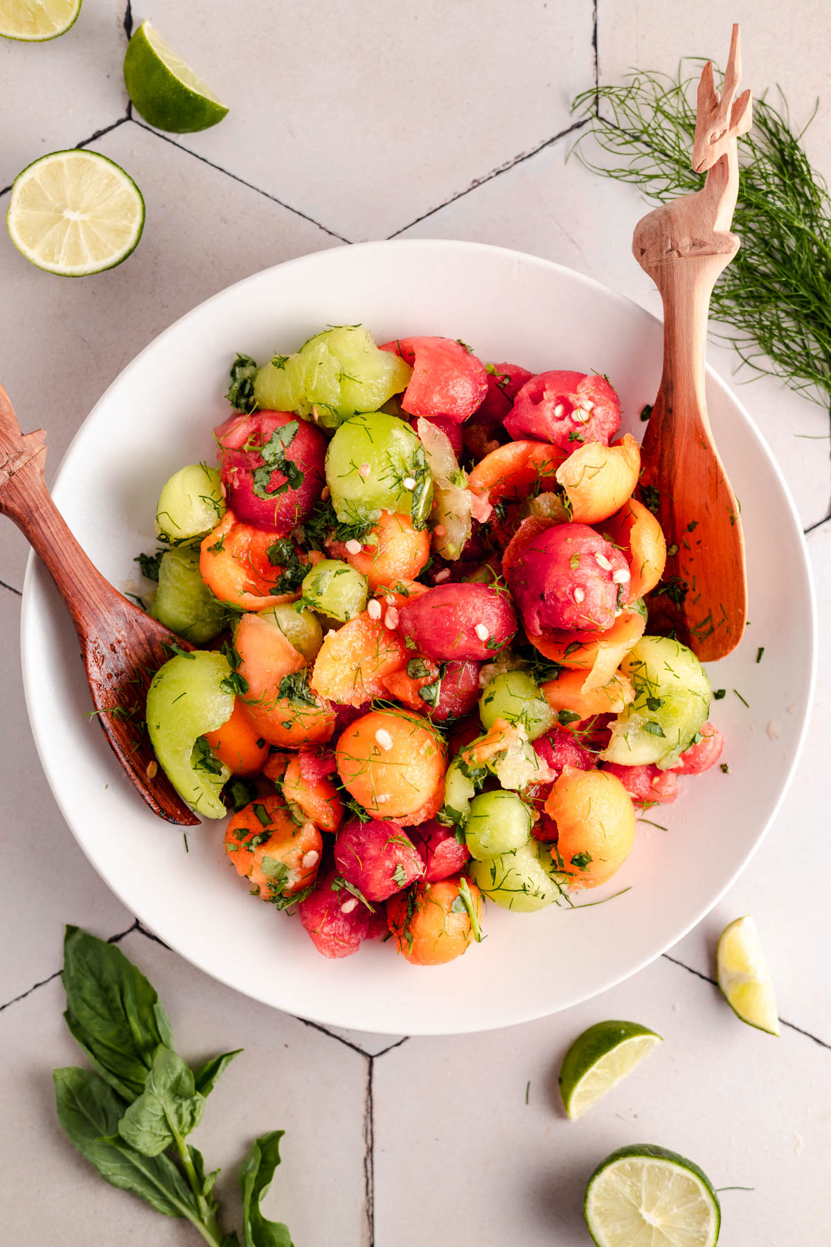 A bowl of mixed melon balls garnished with herbs, with wooden utensils on either side, lime wedges, and herbs surrounding the bowl on a tiled surface.
