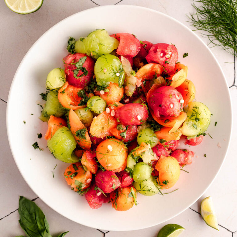 A white plate filled with a colorful melon ball salad including watermelon, cantaloupe, and honeydew, garnished with fresh herbs. Lime wedges and herb leaves are on the table around the bowl.