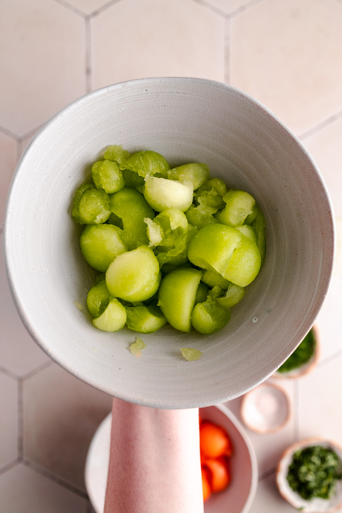 A white bowl containing scooped green melon balls is being held over a tiled surface, with other ingredients slightly blurred in the background.