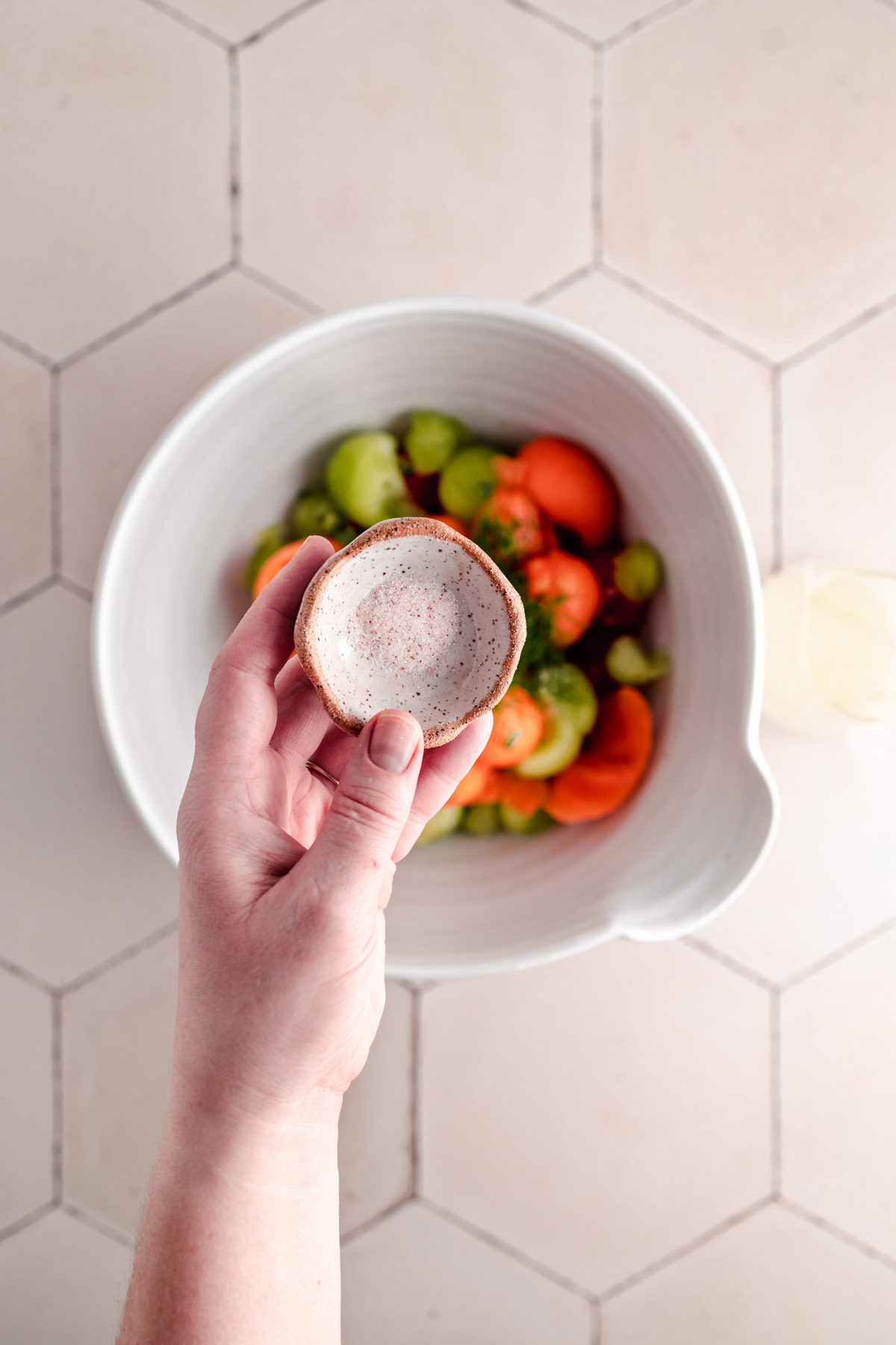 A hand holds a small ceramic bowl above a larger bowl containing tomatoes and other vegetables on a tiled surface.