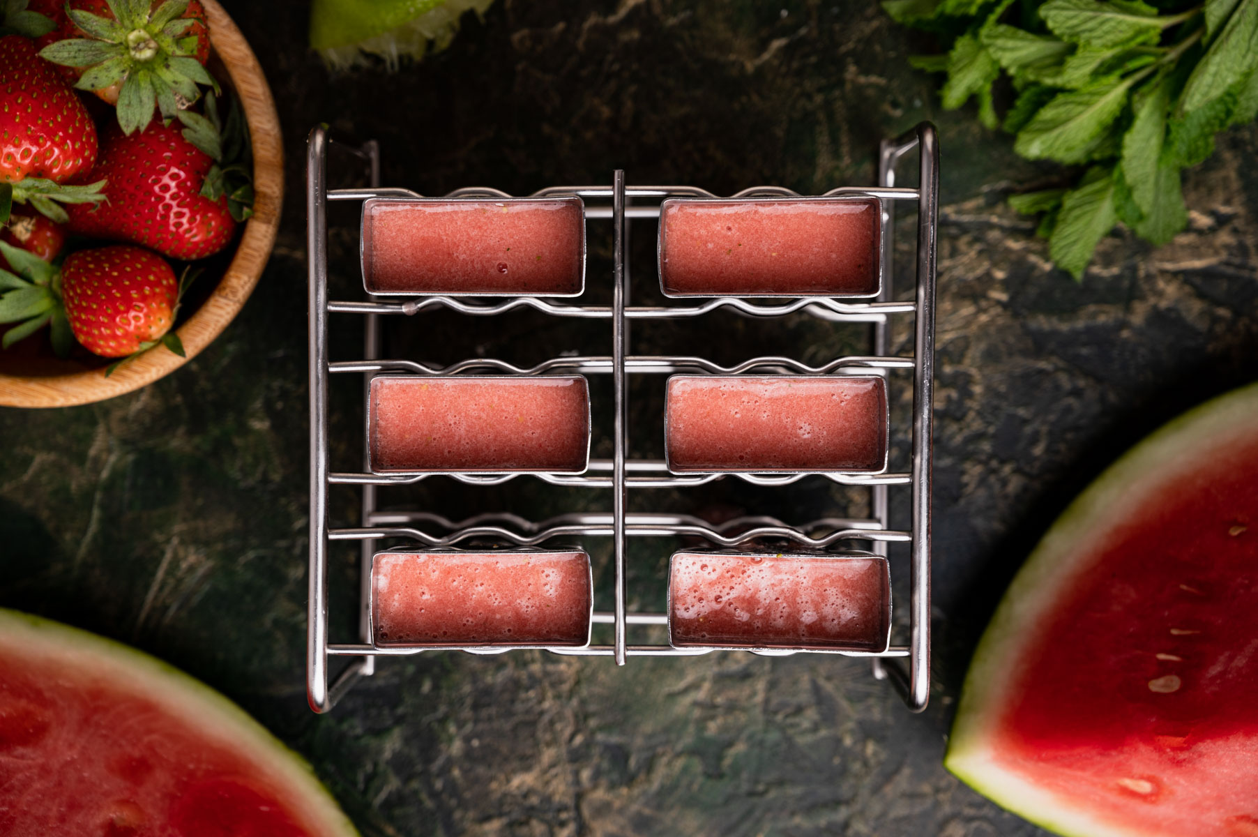 Six strawberry watermelon ice pops in a metal mold, surrounded by fresh strawberries, mint leaves, and watermelon slices.