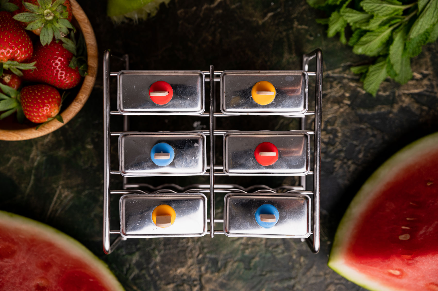 A metal spice rack with six evenly spaced containers, each with a different colored lid, surrounded by strawberries, mint, and watermelon slices on a dark textured surface.