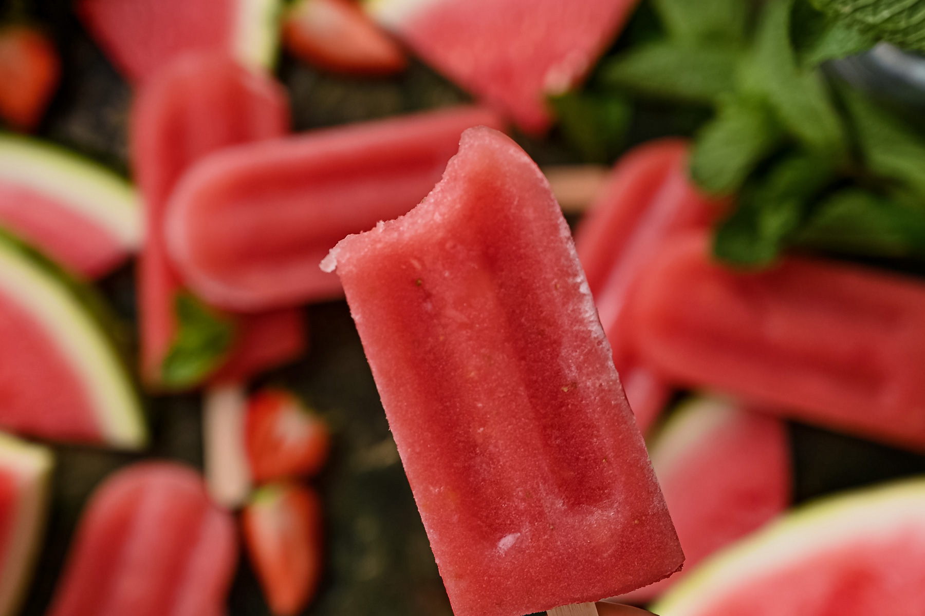 Close-up of a bitten strawberry popsicle held in focus, with whole and sliced strawberries, watermelon slices, and mint leaves blurred in the background.