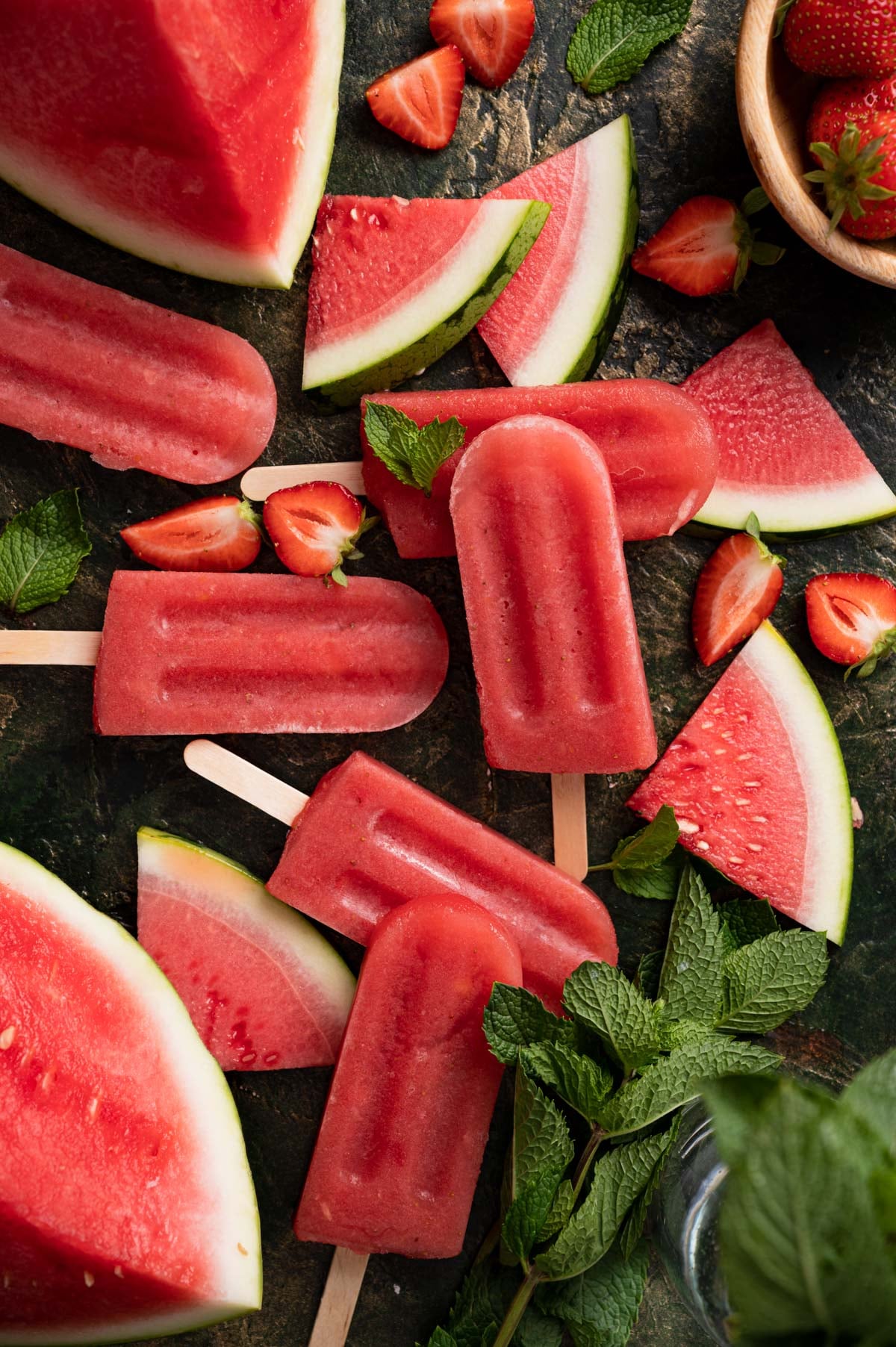 Slices of watermelon, strawberries, mint leaves, and watermelon popsicles are arranged on a dark surface.