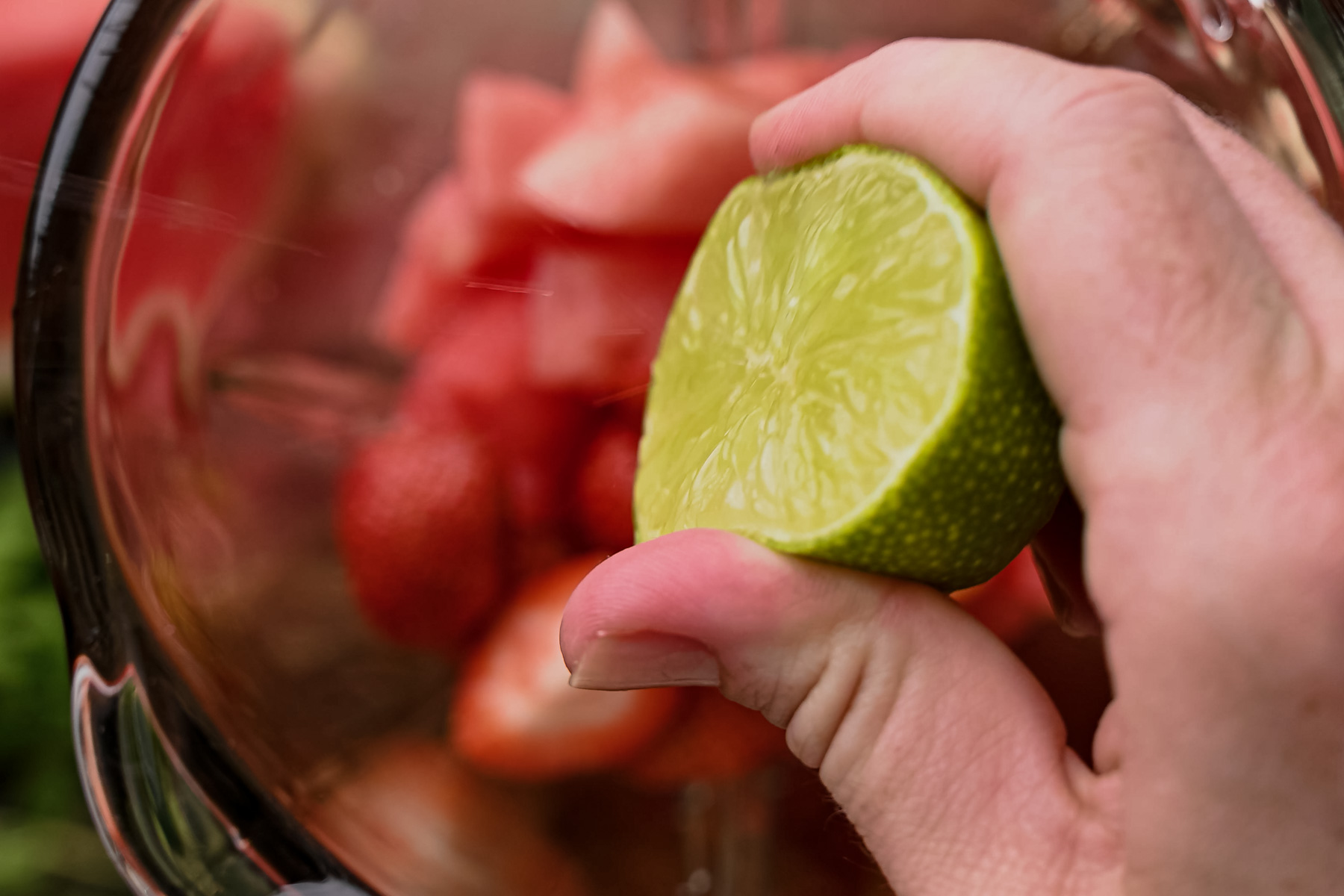 A hand squeezes half a lime into a blender containing cut fruit pieces.