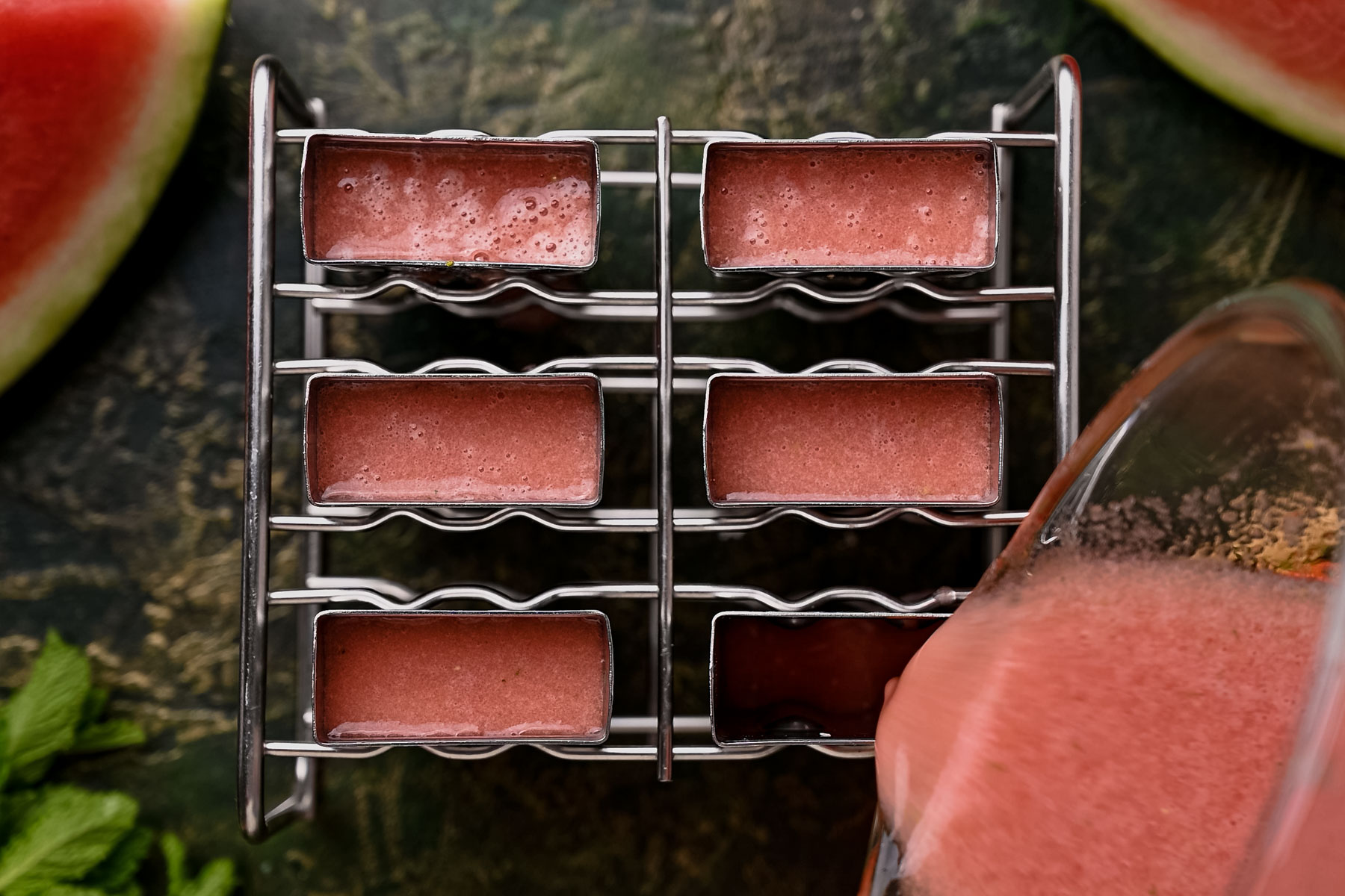 A tray filled with rectangular molds of pink liquid, with additional pink liquid being poured from a pitcher. Green leaves and watermelon slices are visible in the background.