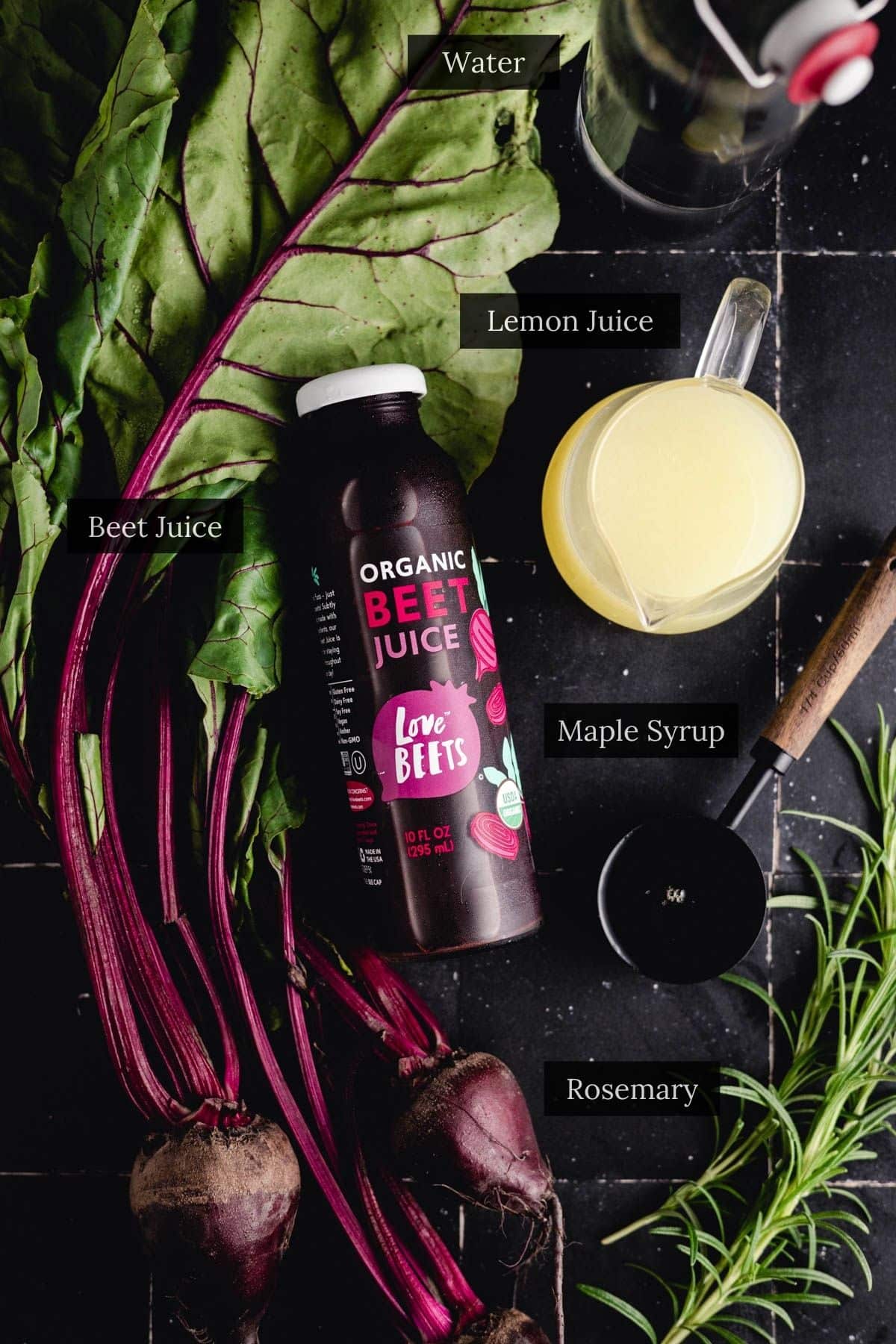 Top-down view of various ingredients labeled as water, lemon juice, organic beet juice, maple syrup, and fresh rosemary arranged on a dark surface with two whole beets and their greens—perfect for crafting an easy beet recipe.