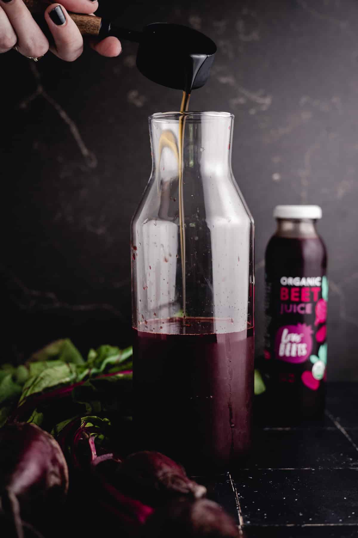 A person pours maple syrup into a clear glass bottle with beet juice inside. A beet juice bottle labeled "Organic Beet Juice" is in the background, along with fresh beets and greens on a dark surface.