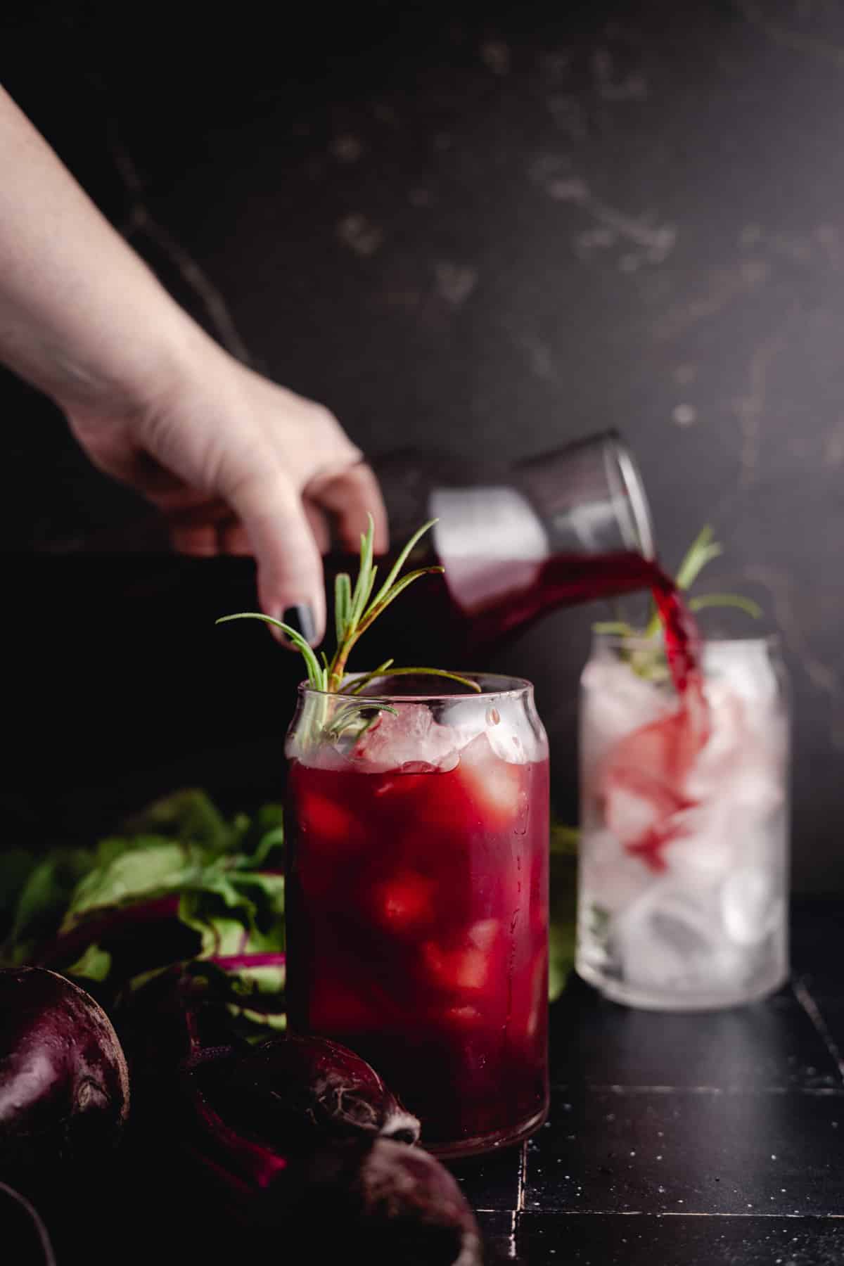 A hand pouring beet lemonade into a glass filled with ice. Another glass with a similar drink sits nearby. Fresh herbs are used as garnish, and beets are visible in the foreground.