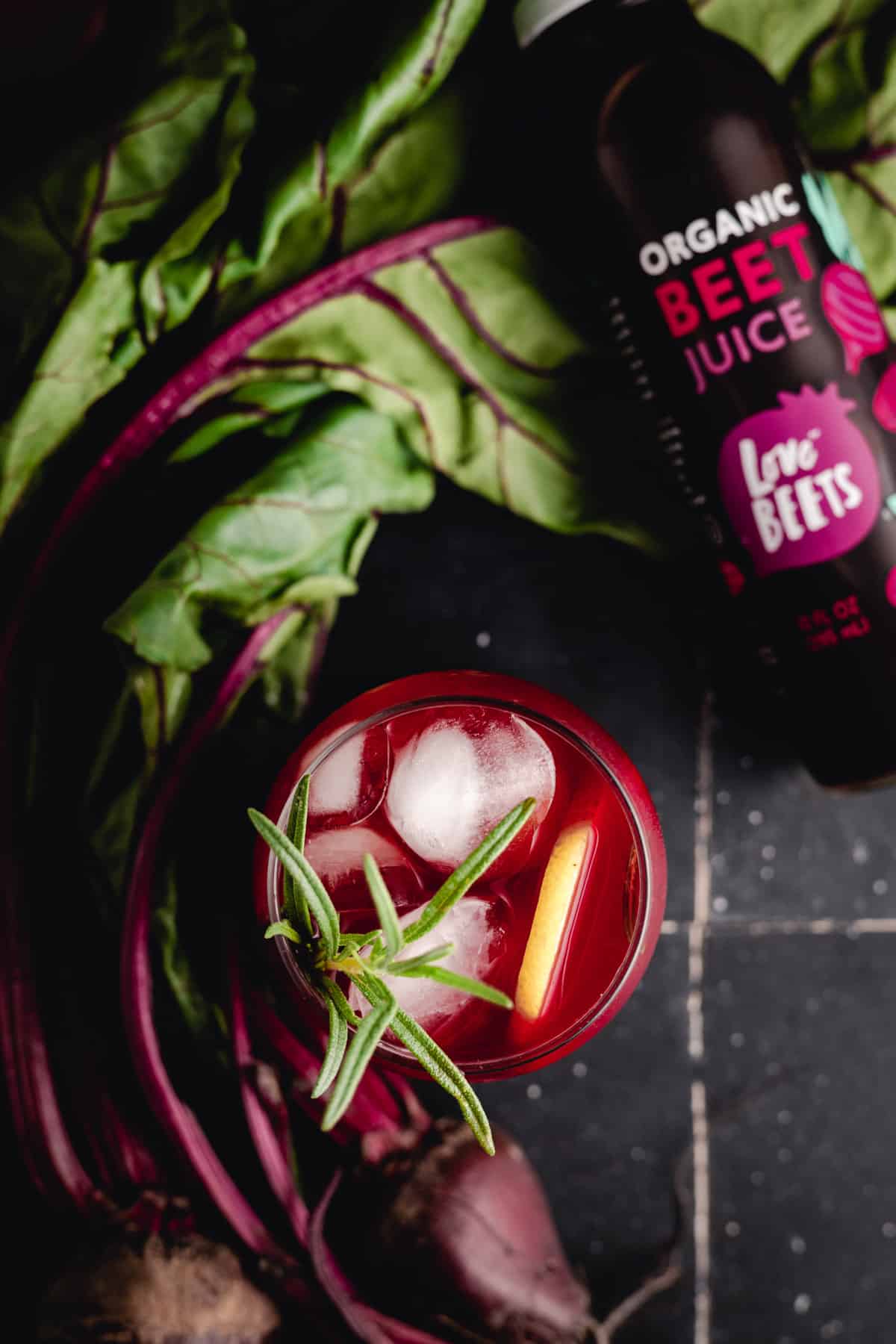 Top view of a glass of iced beet lemonade garnished with a sprig of rosemary and a lemon peel next to a bottle of organic beet juice and fresh beet roots on a dark surface.