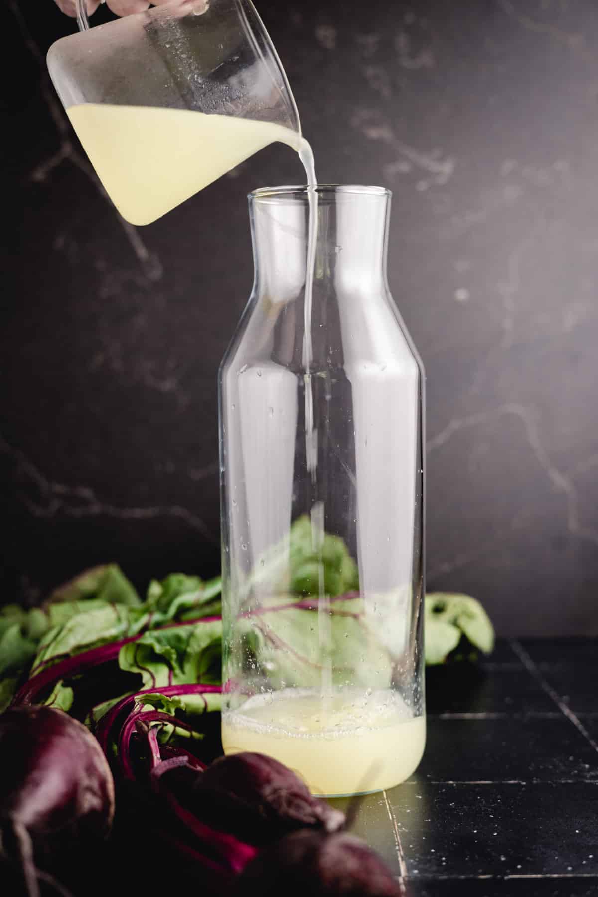 A hand pours lemon juice from a glass container into an empty glass jug, with beetroot and greens in the background.