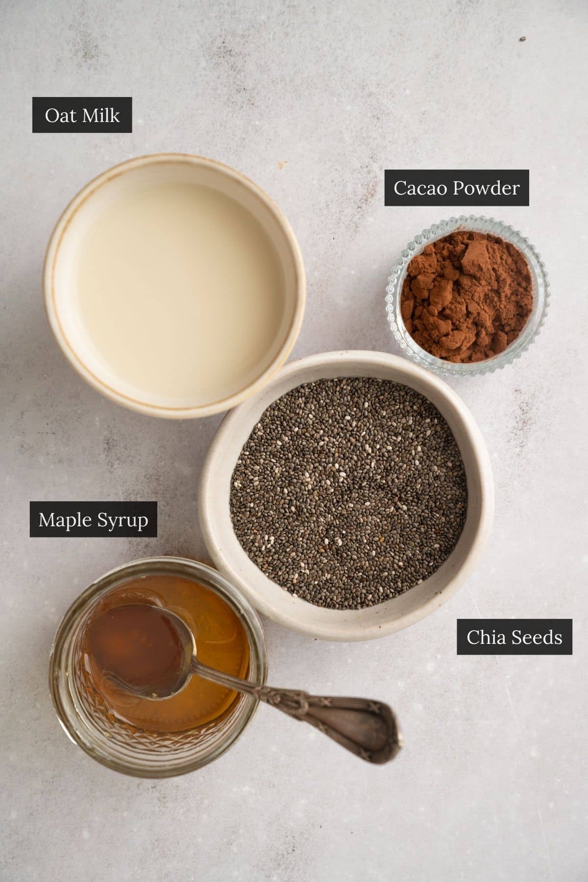 Top view of ingredients for chocolate chia pudding, including oat milk, cacao powder, chia seeds, and maple syrup, each in separate bowls labeled with black text.