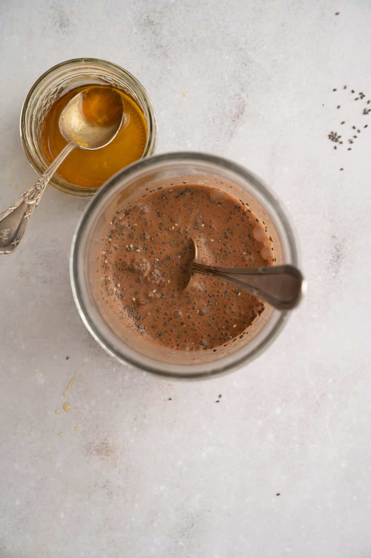 A glass jar filled with a brown mixture and a spoon inside, next to a jar of maple syrup with a spoon on the edge. Specks of black and maple syrup drops on the white surface.