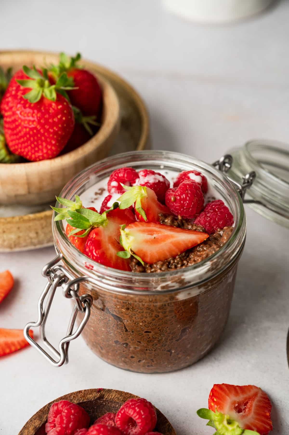 A jar of chocolate chia pudding topped with fresh strawberries and raspberries is placed on a table. A bowl of strawberries and a small plate of raspberries are in the background.
