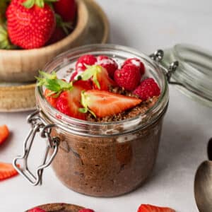 A glass jar filled with chocolate chia pudding topped with strawberries and raspberries, surrounded by sliced strawberries and wooden spoons on a table.