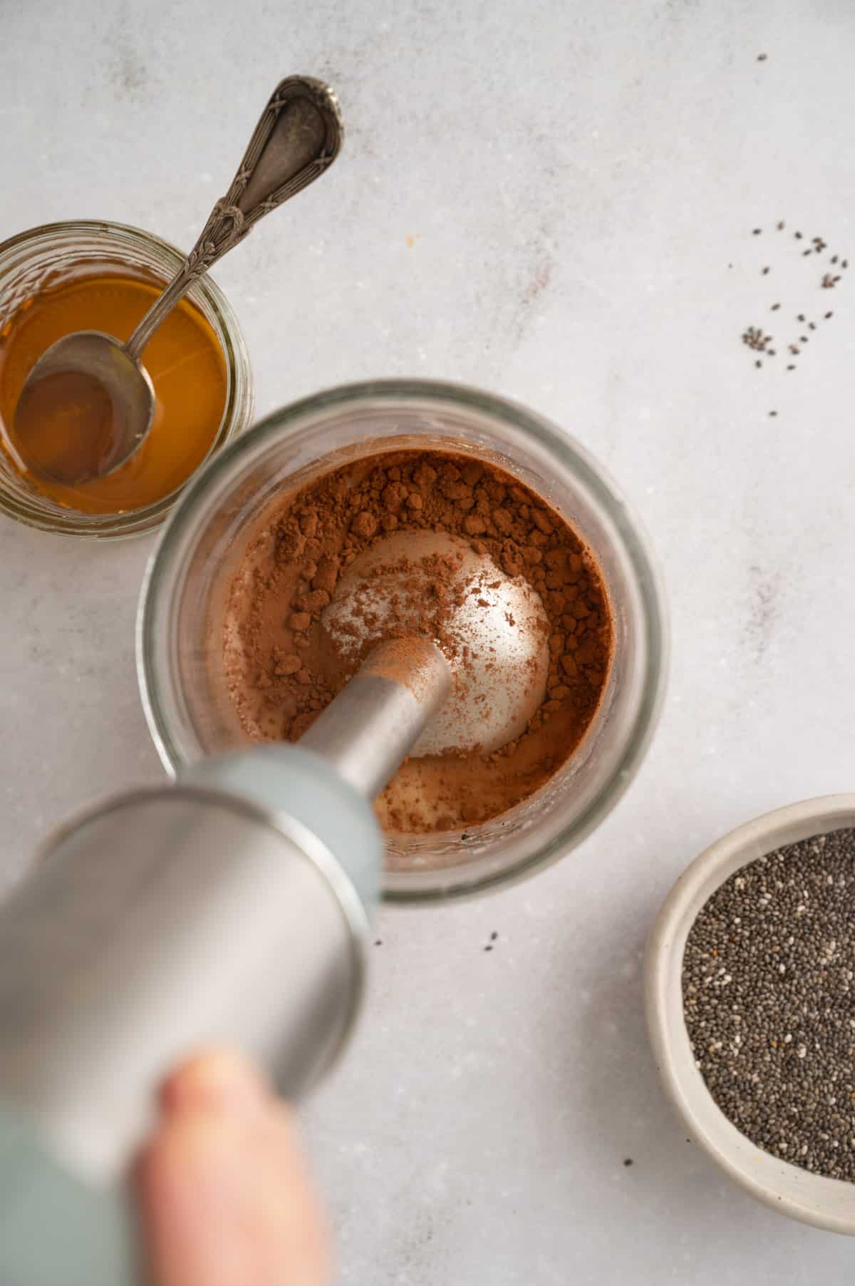 A hand using an immersion blender mixes cocoa powder in a glass container. Nearby are a jar of maple syrup with a spoon and a bowl of chia seeds on a white surface.