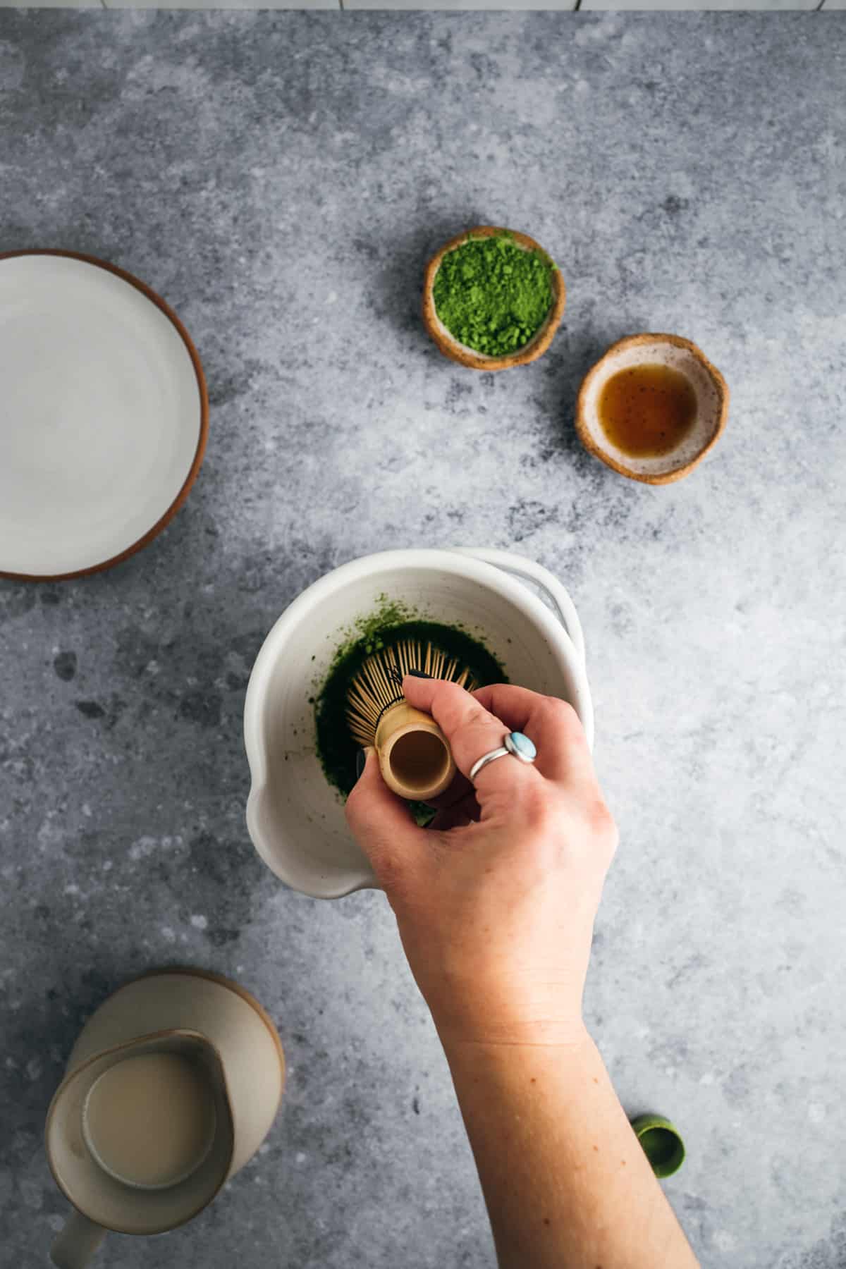 A person prepares matcha tea by whisking green matcha powder with a bamboo whisk in a white bowl. Nearby are small bowls containing matcha powder and another ingredient, a jug, and an empty plate.
