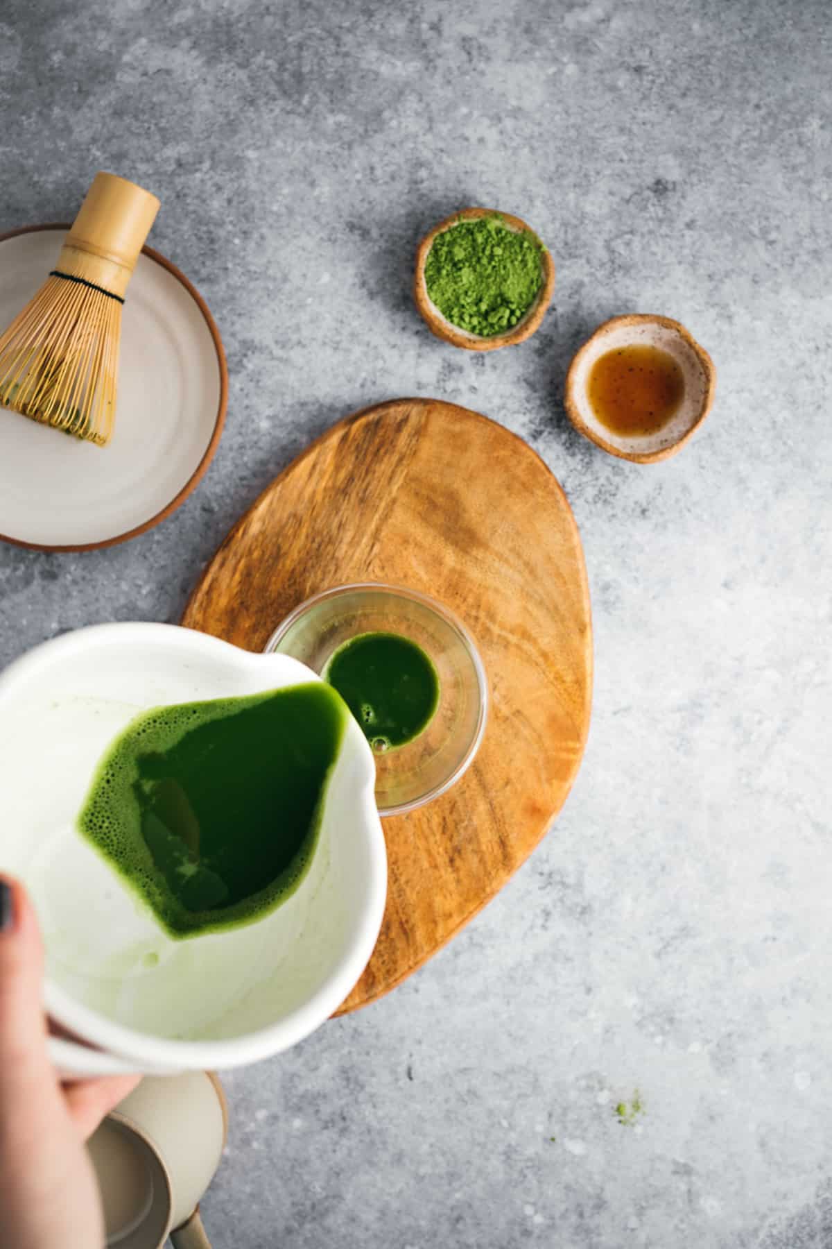 A person pours green matcha tea from a white bowl into a glass on a wooden board, with matcha powder and a whisk nearby on a gray surface.