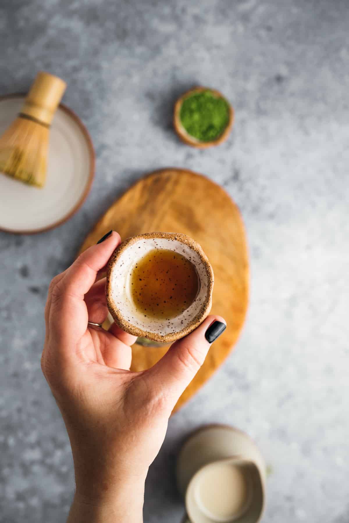 A hand holds a small, rustic ceramic cup with matcha tea. A bamboo whisk, a bowl of matcha powder, and a wooden board are in the background.