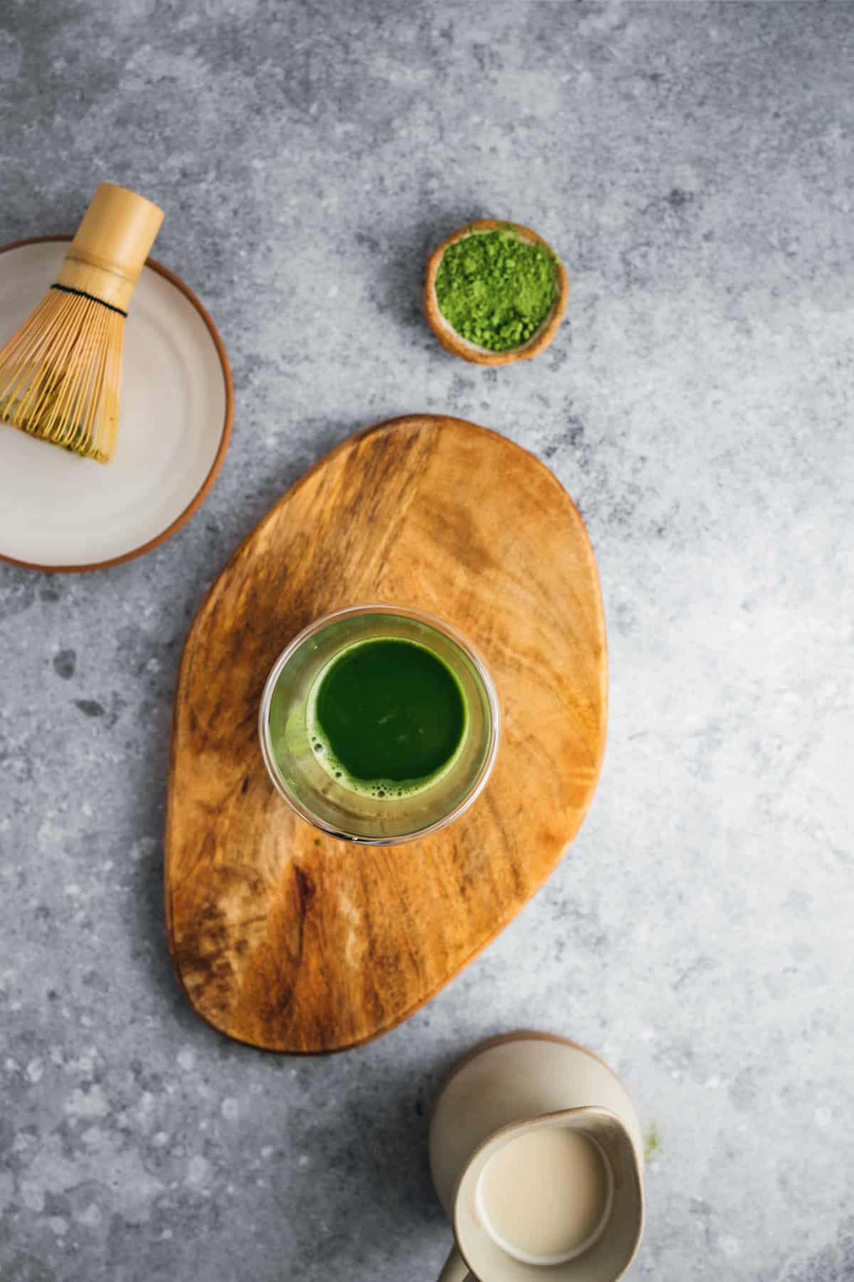 Top view of a freshly made matcha green tea on a wooden board, with a whisk and a bowl of matcha powder nearby.