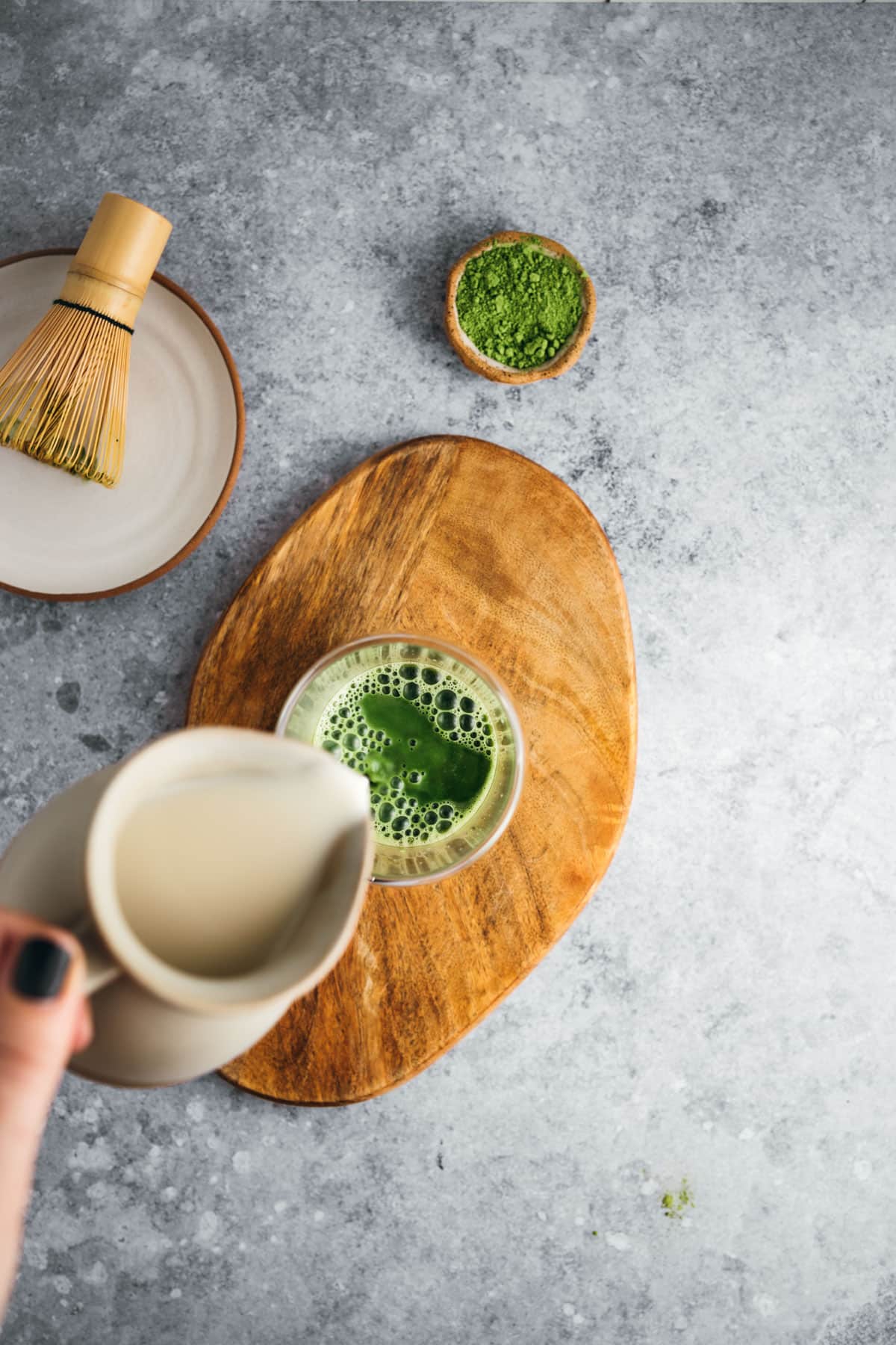 A hand pours milk from a pitcher into a glass of green matcha on a wooden board. A bowl of matcha powder and a bamboo whisk are beside the glass.
