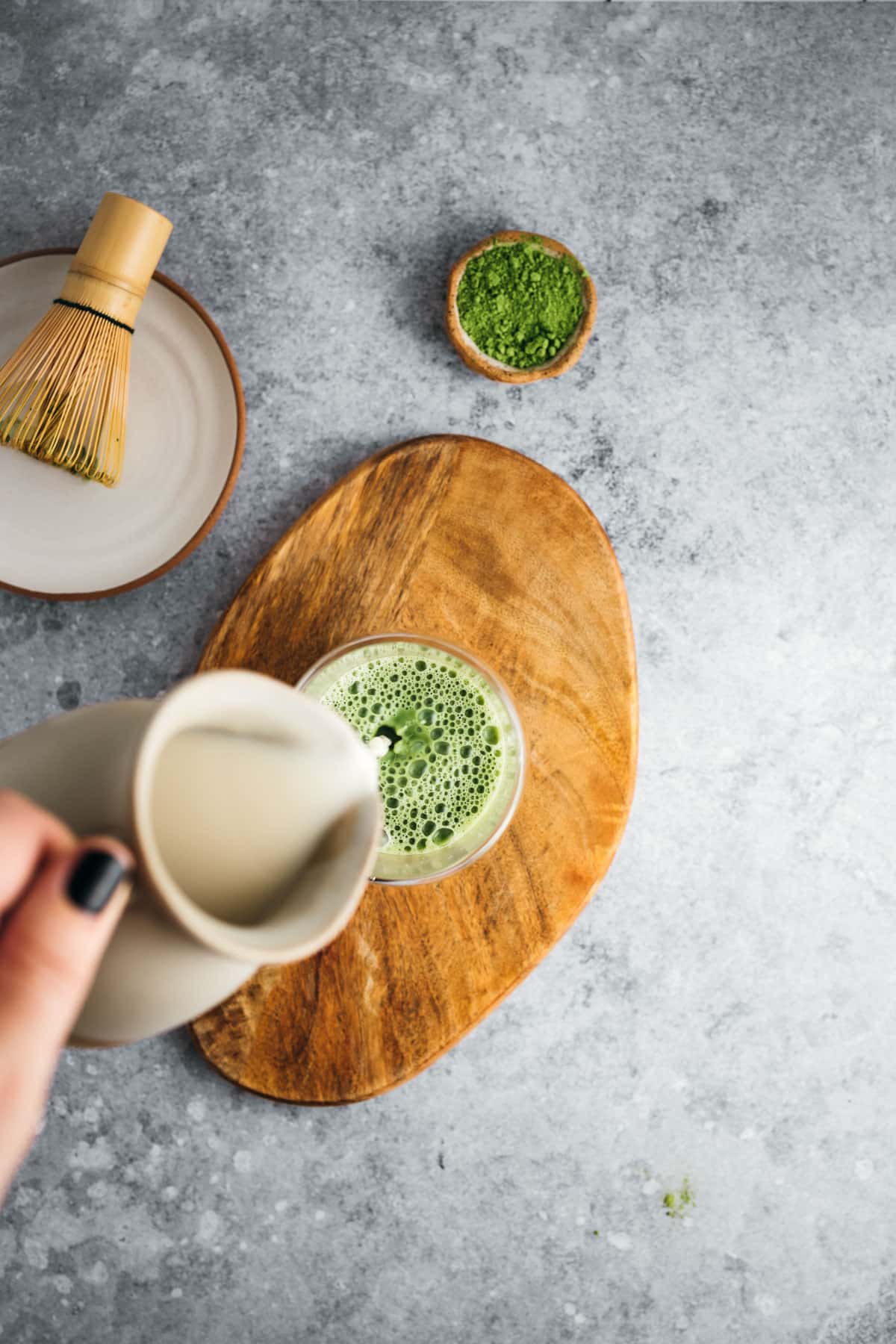 A hand pours milk into a glass of matcha green tea on a wooden board. A bamboo whisk and a small bowl of matcha powder are nearby on a gray countertop.
