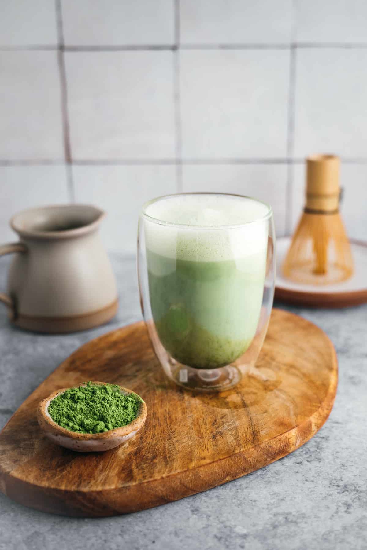 A glass of matcha latte sits on a wooden board with a small bowl of matcha powder, a whisk, and a ceramic pitcher in the background.