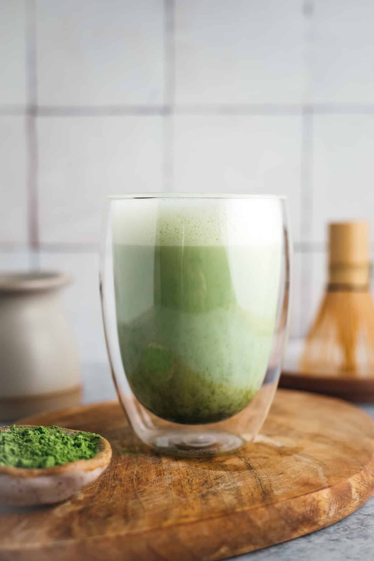 A glass cup of matcha latte on a wooden board, with a small bowl of matcha powder and a bamboo whisk in the background.