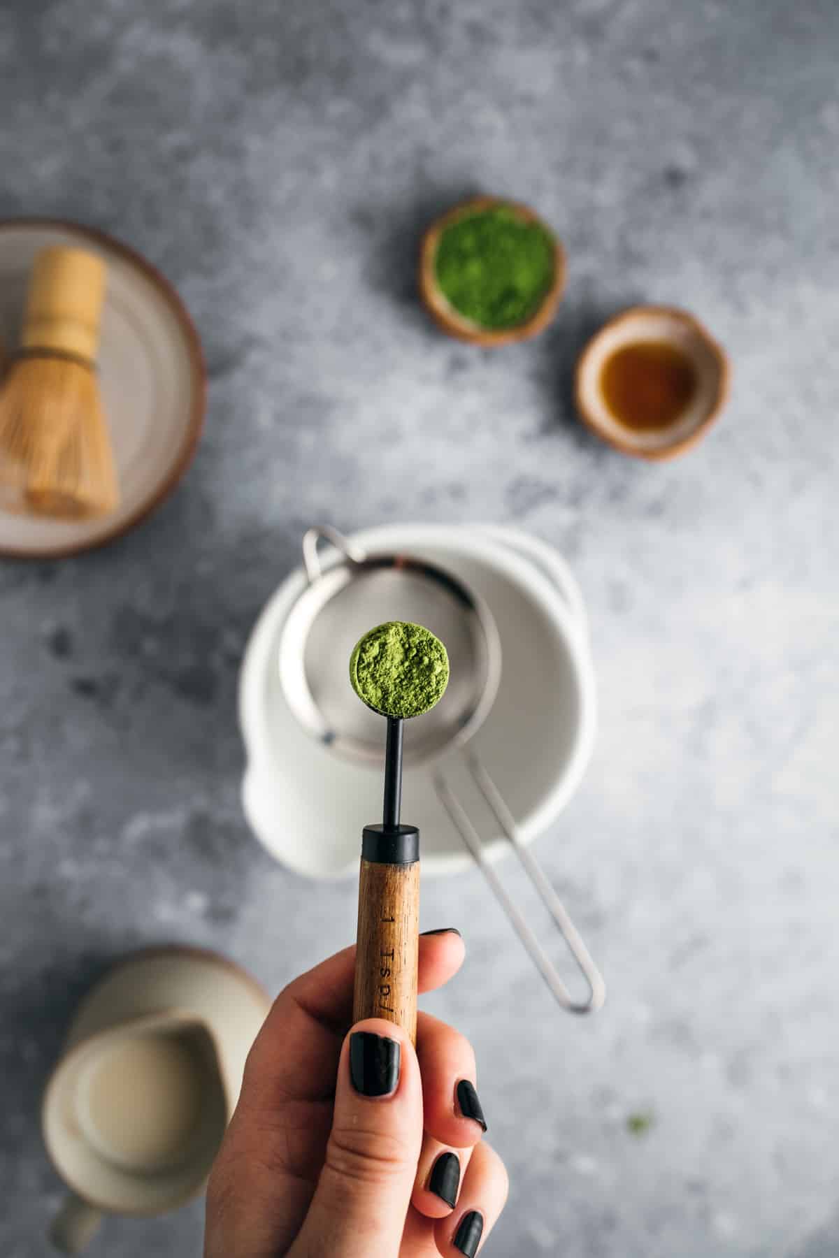 Hand holding a spoon with green matcha powder over a tea strainer and bowl. In the background, there are bowls with more matcha powder and a bamboo whisk.