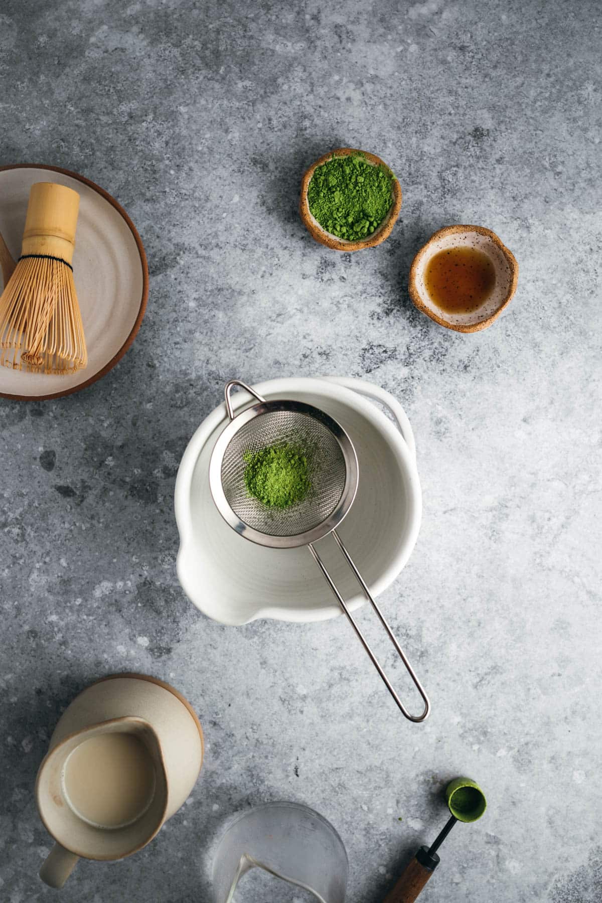Overhead view of a matcha tea preparation setup on a grey countertop, featuring a cup, sieve with matcha powder, whisk, bowl, small dishes of matcha powder and tea, and a bowl of milk.