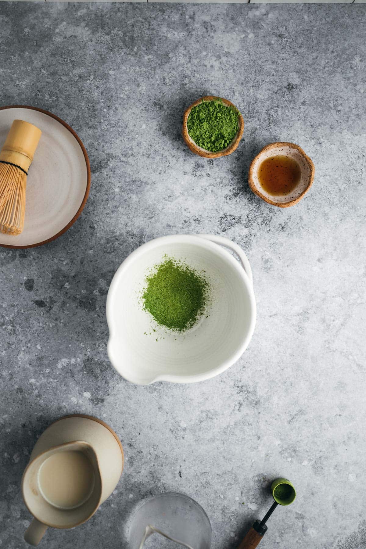 A ceramic bowl containing matcha powder, a whisk, a small wooden bowl with more matcha, a ceramic cup with liquid, and a spoon with matcha powder are arranged on a gray surface.