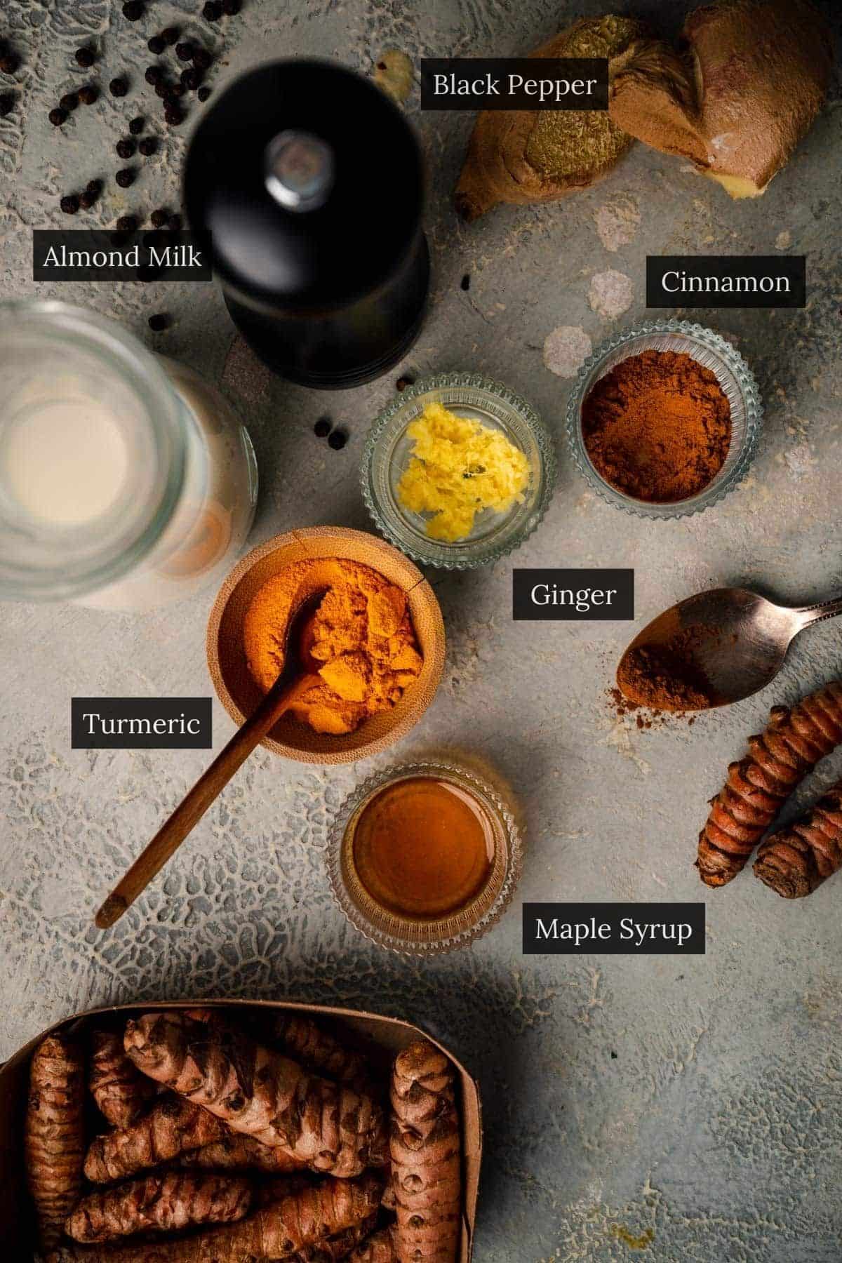 A top-down view of turmeric latte ingredients, including almond milk, black pepper, ginger, cinnamon, turmeric, and maple syrup, arranged beautifully on a textured surface.