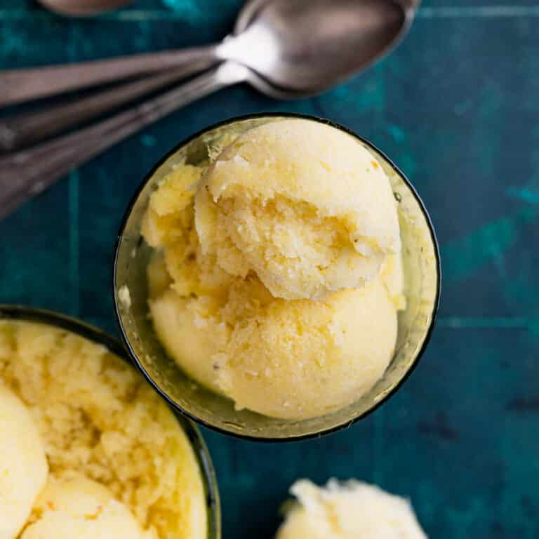 A glass bowl filled with scoops of yellow sorbet, placed on a dark green surface. Three metal spoons are visible in the background.