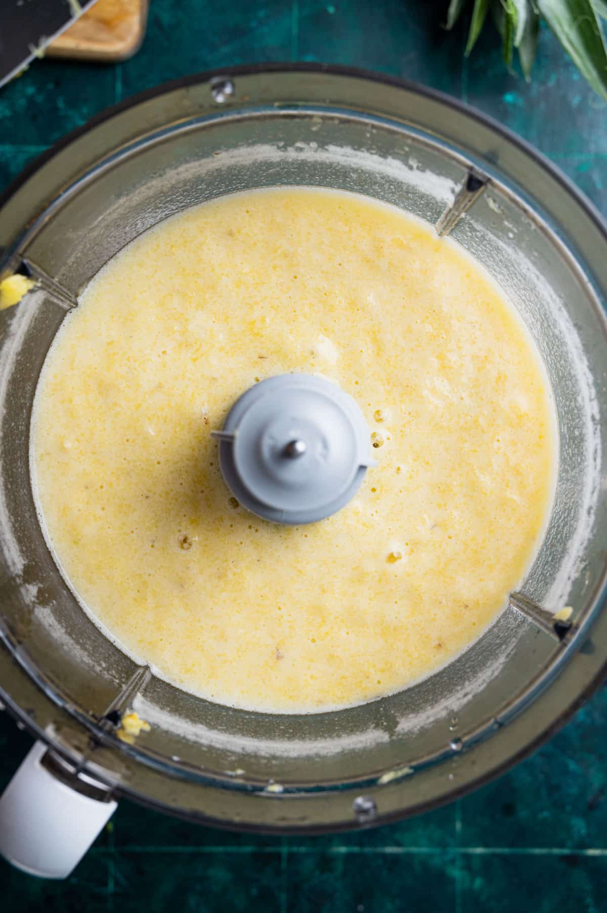 A food processor containing a yellow, frothy mixture viewed from above on a dark green surface.