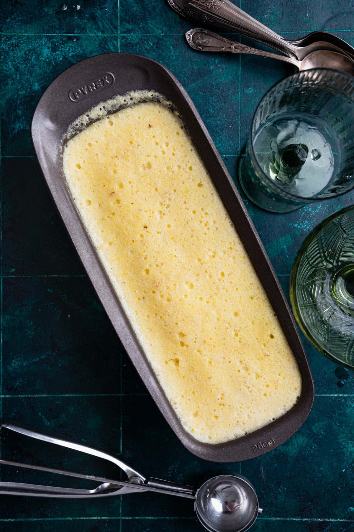 A rectangular baking dish filled with a baked custard-like dish, surrounded by glasses and metal utensils on a dark, textured surface.