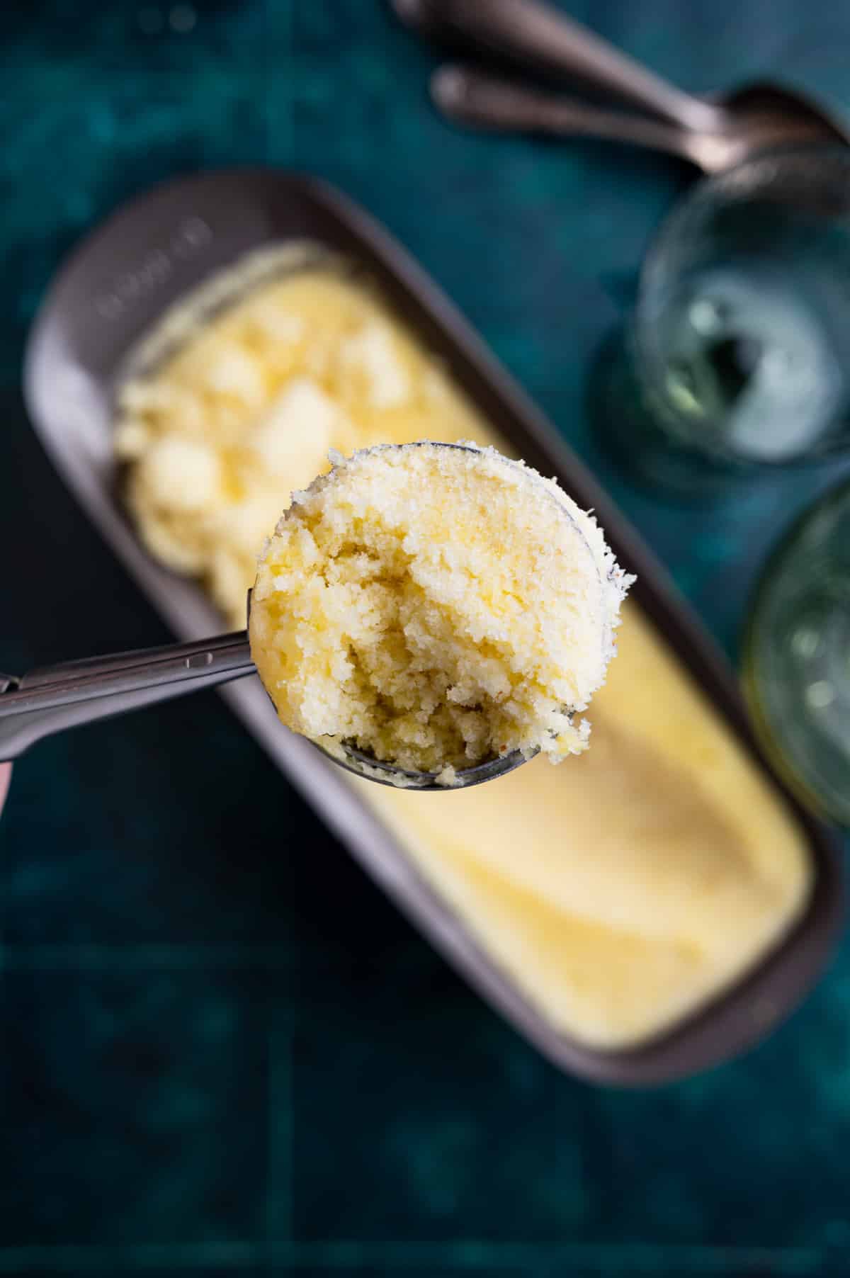 A close-up of a spoon scooping out a portion of homemade ice cream from a rectangular container placed on a teal surface, with two empty glasses and spoons in the background.