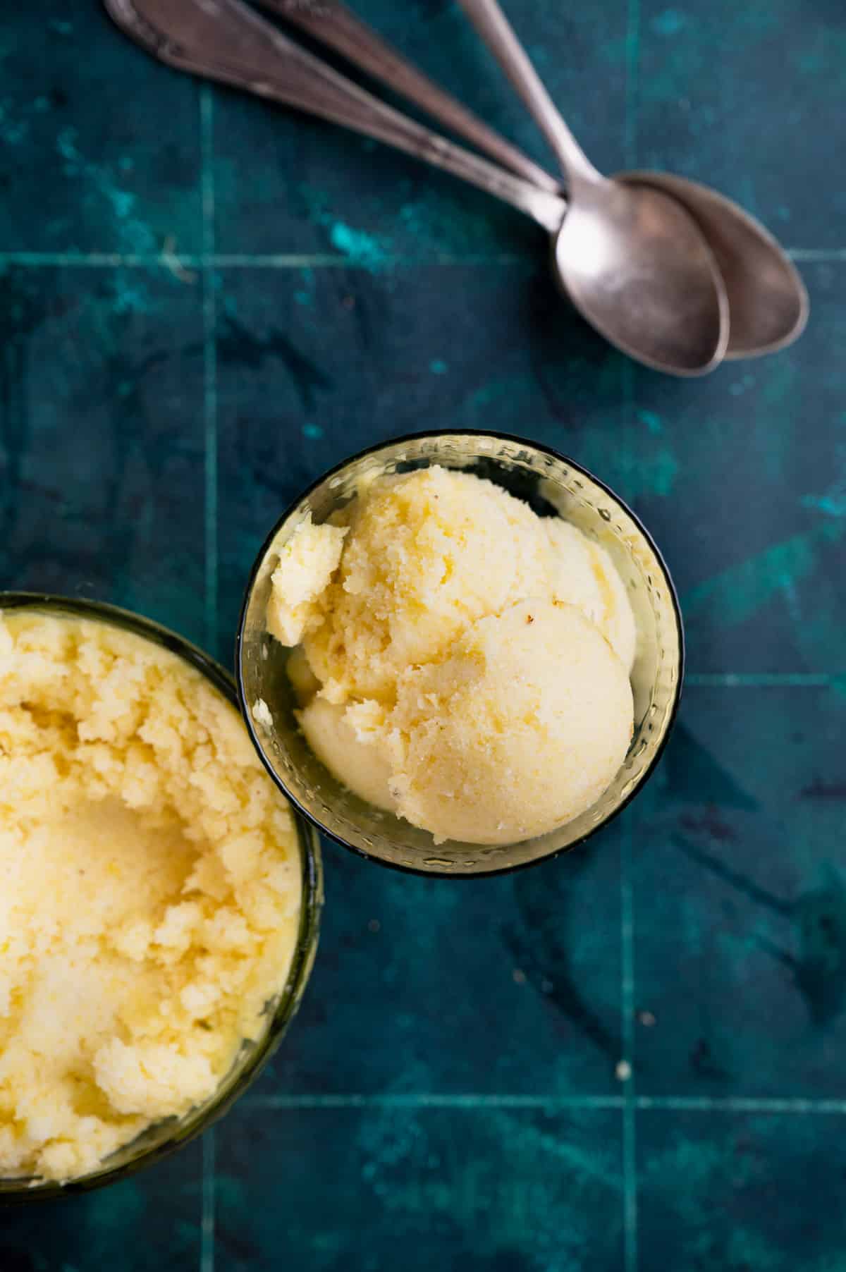 Top view of scoops of yellow ice cream in a glass bowl on a dark green textured surface, with two silver spoons placed nearby.