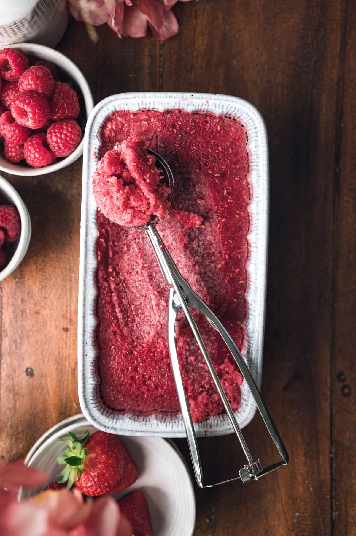 A rectangular dish filled with raspberry sorbet, an ice cream scoop resting on top. Surrounding the dish are bowls holding fresh raspberries and strawberries on a wooden surface.