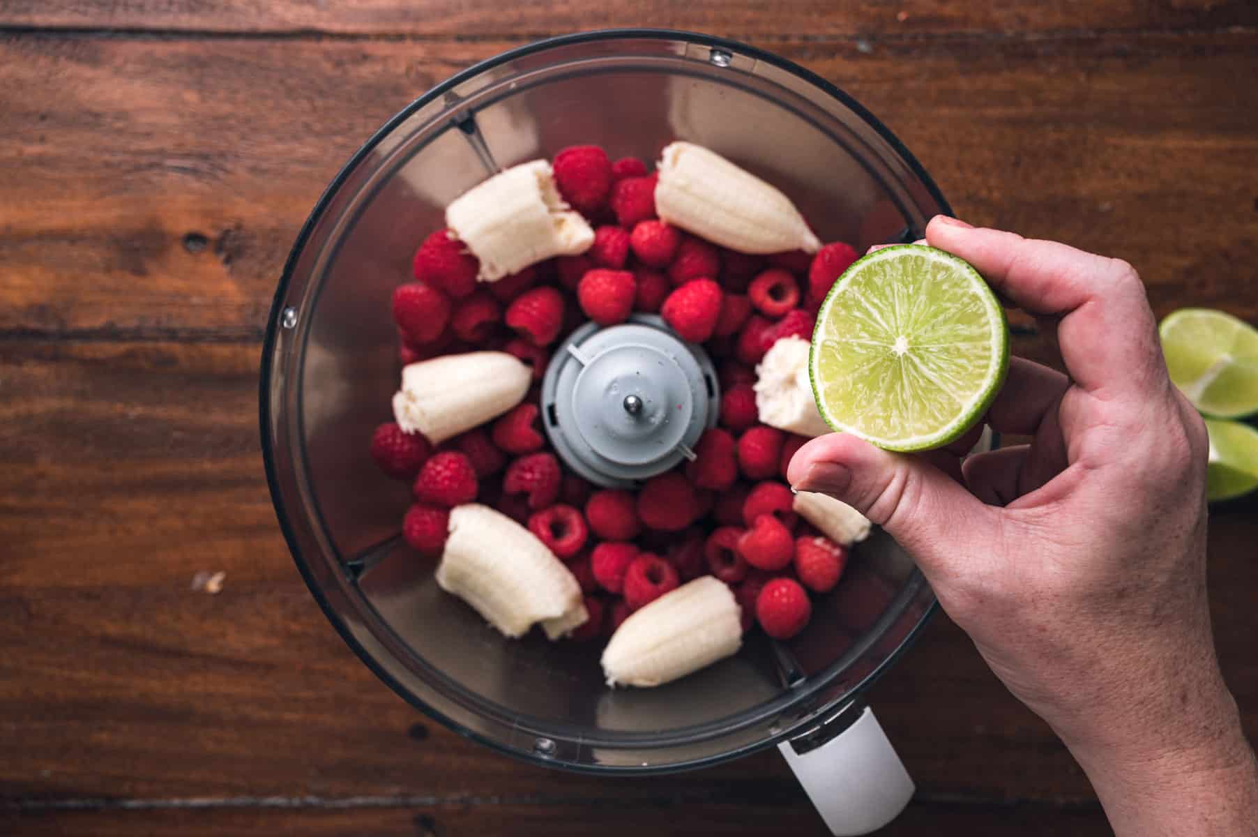 A hand holds a halved lime above a food processor containing bananas and raspberries on a wooden surface.