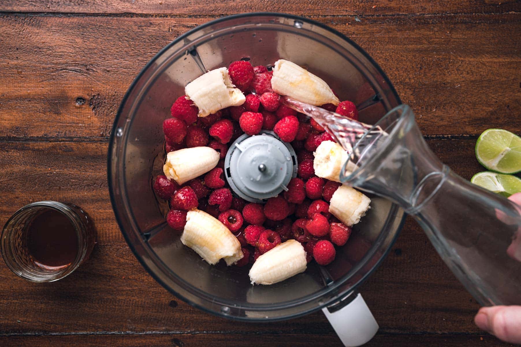 A food processor bowl containing raspberries and banana pieces has water being poured into it. A small cup of liquid and lime halves are nearby on a wooden surface.