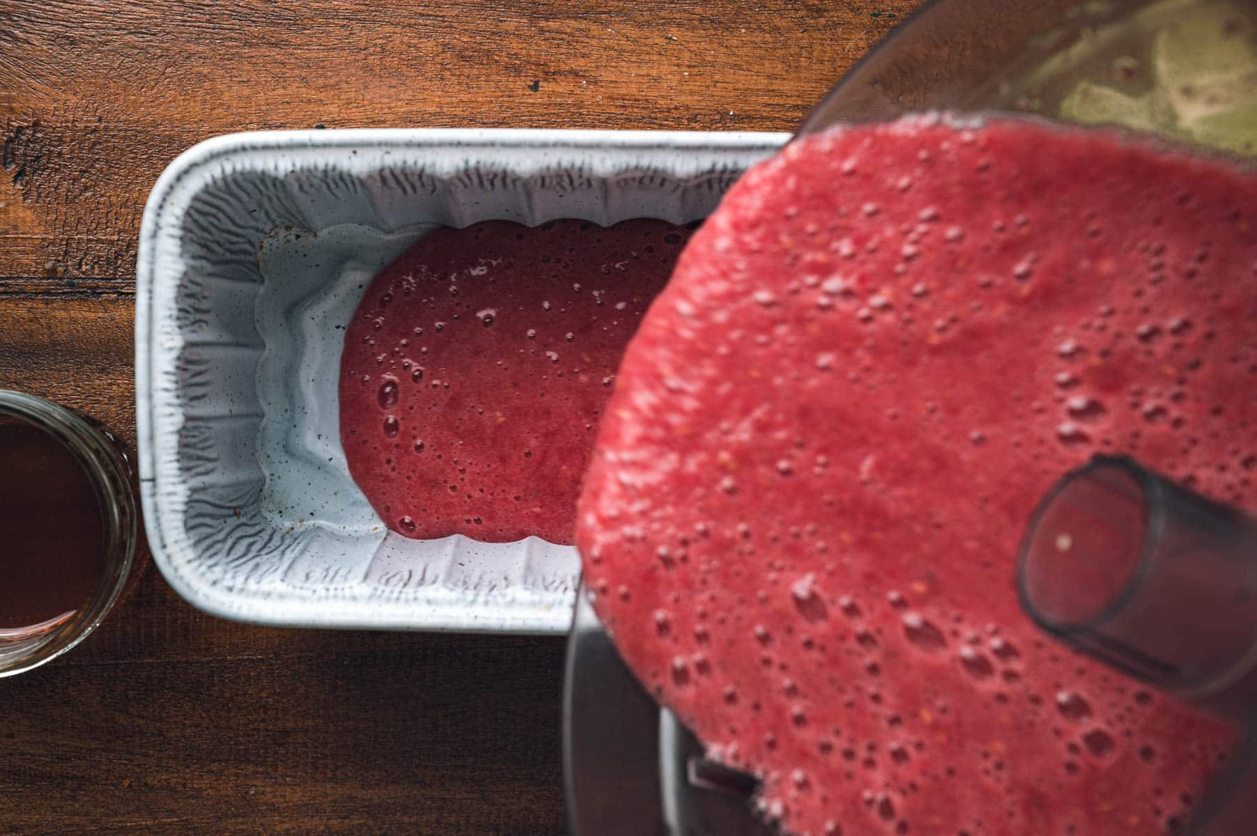 A blender is pouring raspberry sorbet mixture into a rectangular baking dish on a wooden surface, with a glass of dark liquid nearby.